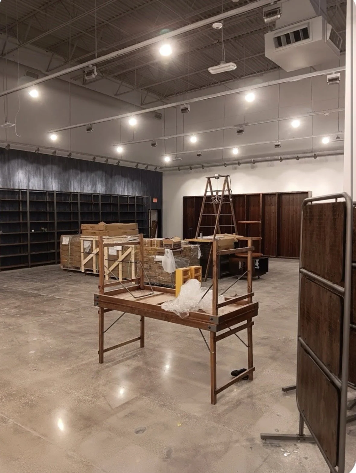 Industrial warehouse interior with wooden display fixtures, packed crates, shelving, and a ladder under bright ceiling lights