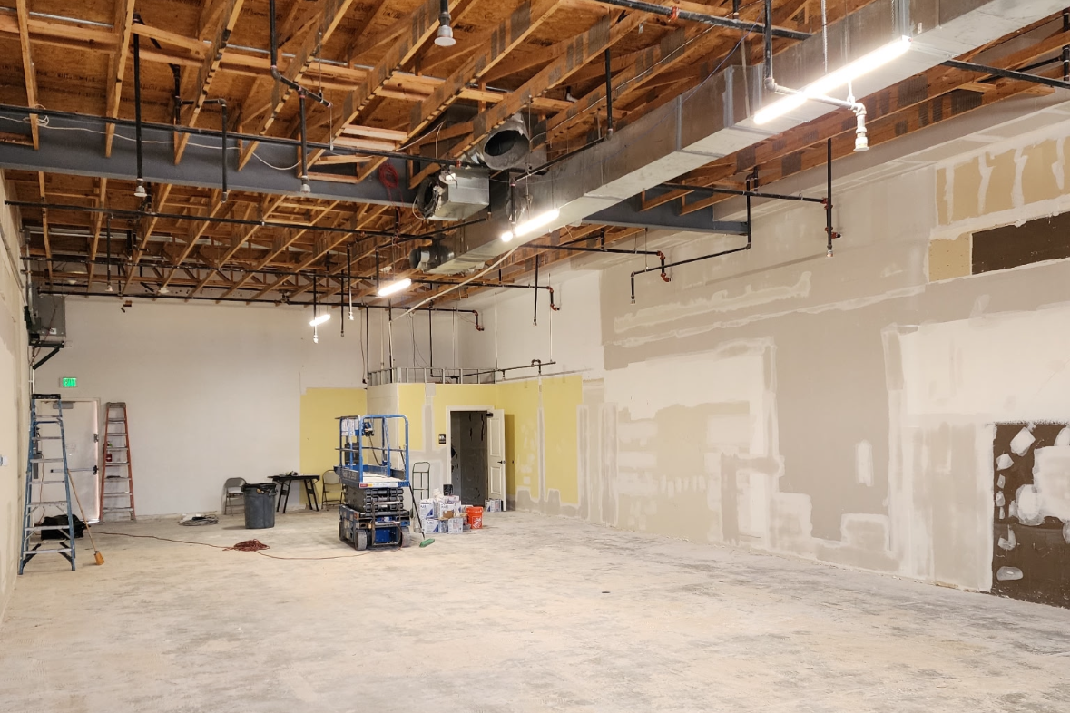 Commercial interior under construction with exposed ceiling, drywall repairs, electrical and plumbing lines, ladders, paint supplies, and a lift.