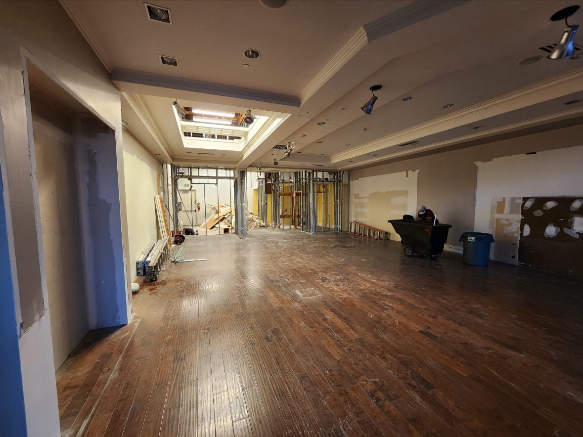 Large unfinished commercial interior under renovation with exposed wall framing, construction materials, utility cart, and worn hardwood floor