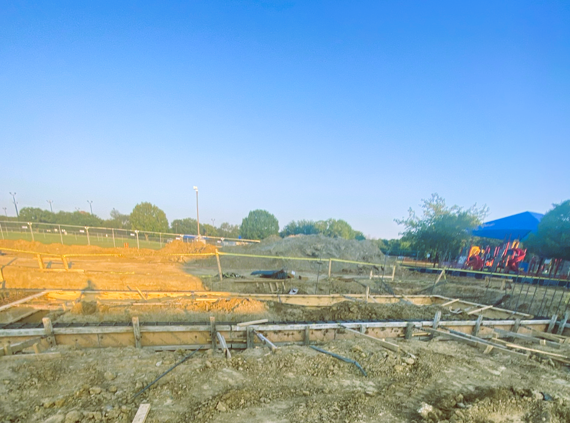 Construction site with wooden foundation forms, exposed dirt, fencing, trees, and a playground under a clear blue sky