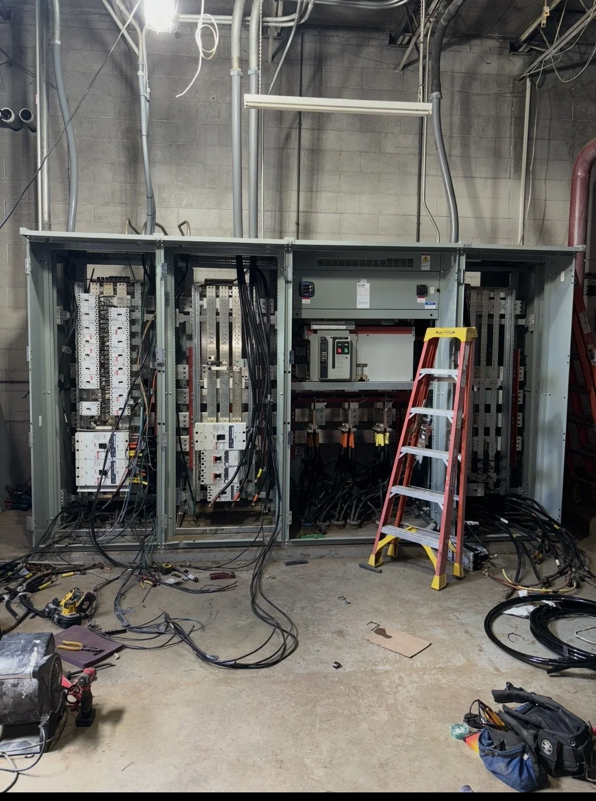 Open electrical switchgear cabinets with circuit breakers, wiring, and cables in an industrial utility room, alongside a red step ladder.