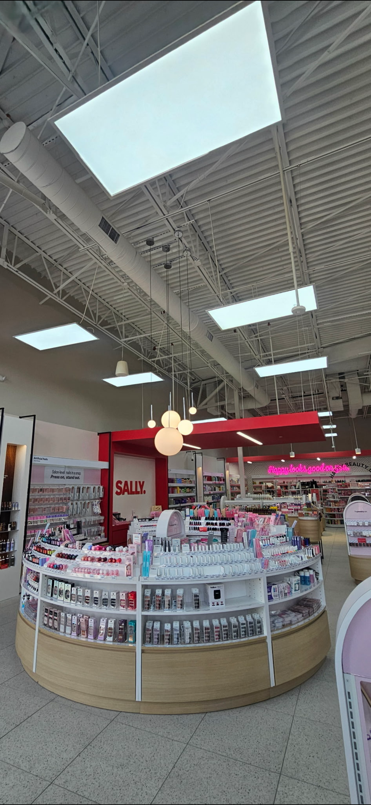 Interior of a Sally Beauty store with shelves displaying nail polish, artificial nails, and beauty products beneath an industrial ceiling with skylights.