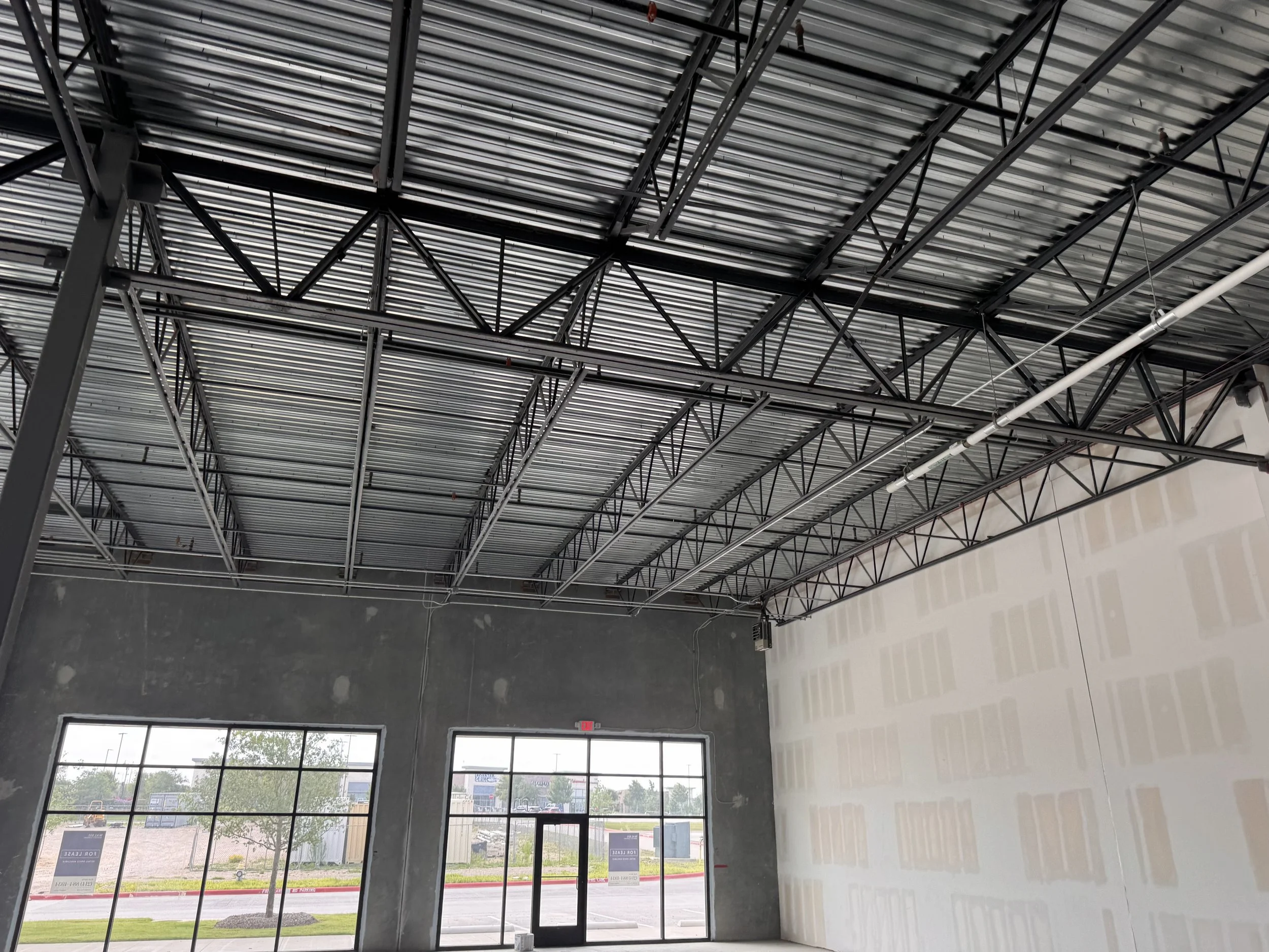 Interior of a commercial building under construction with exposed steel roof trusses, corrugated metal ceiling, plumbing, unfinished drywall, and large storefront windows.