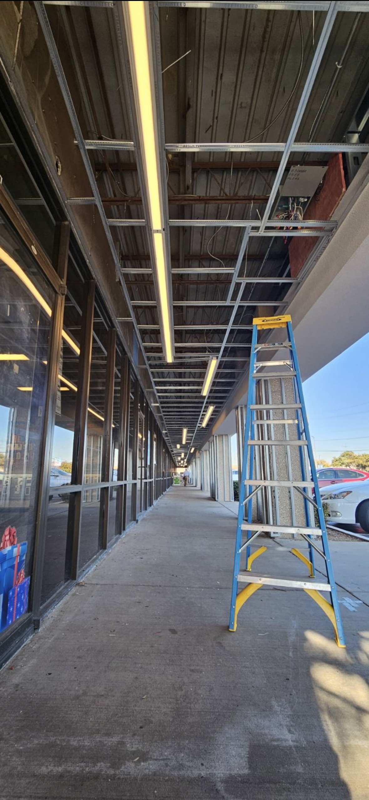 Commercial building walkway under construction with exposed ceiling, fluorescent lights, and a blue step ladder.