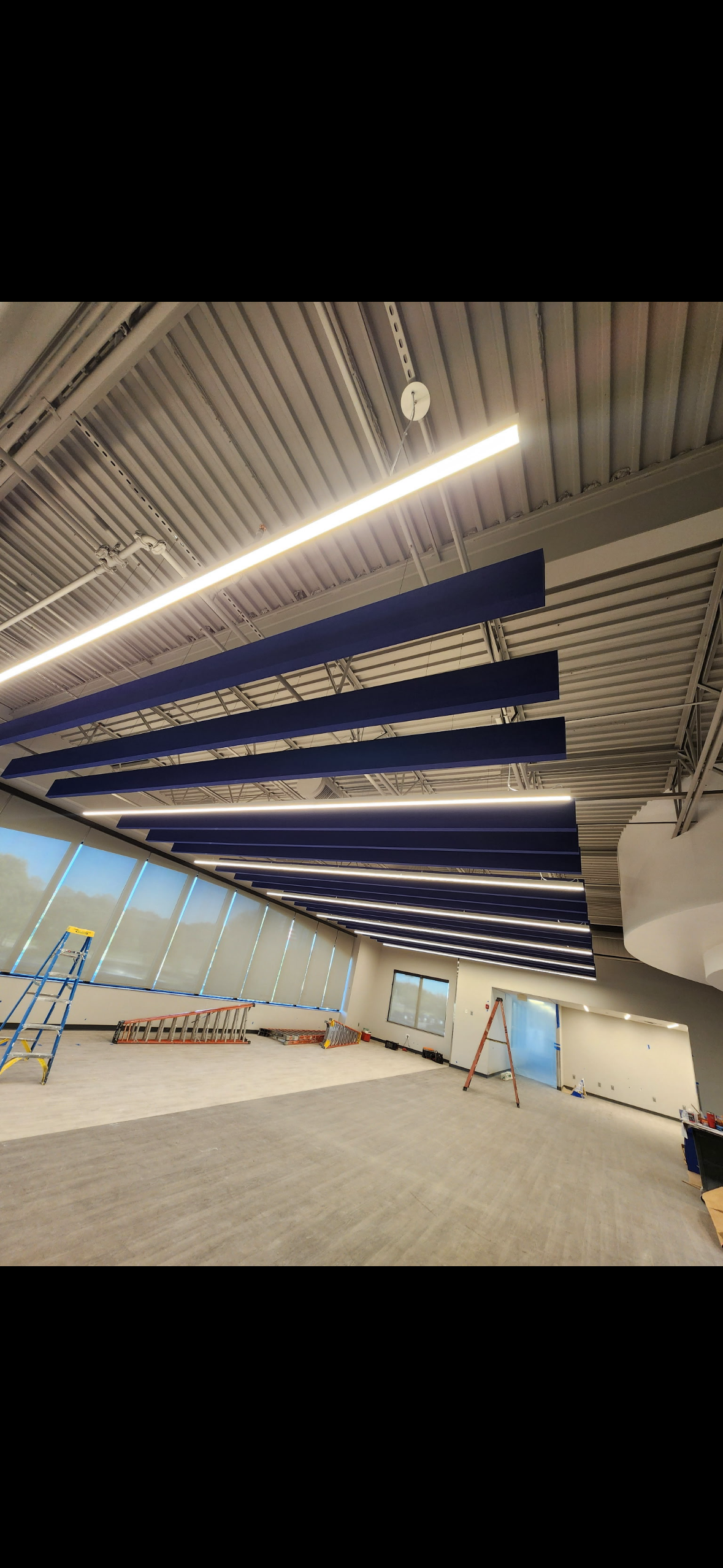 Unfinished commercial interior with blue suspended ceiling panels, fluorescent lights, windows, ladders, and construction equipment.