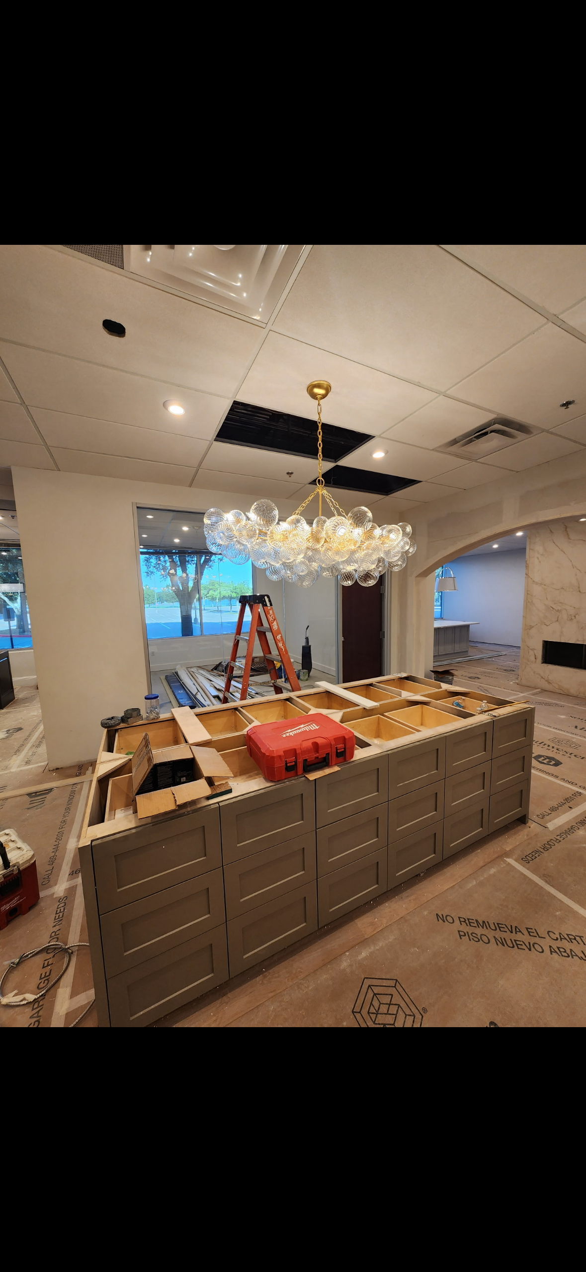 Commercial interior under renovation with a large glass-ball chandelier, unfinished reception desk, ladder, tools, and exposed ceiling panels.