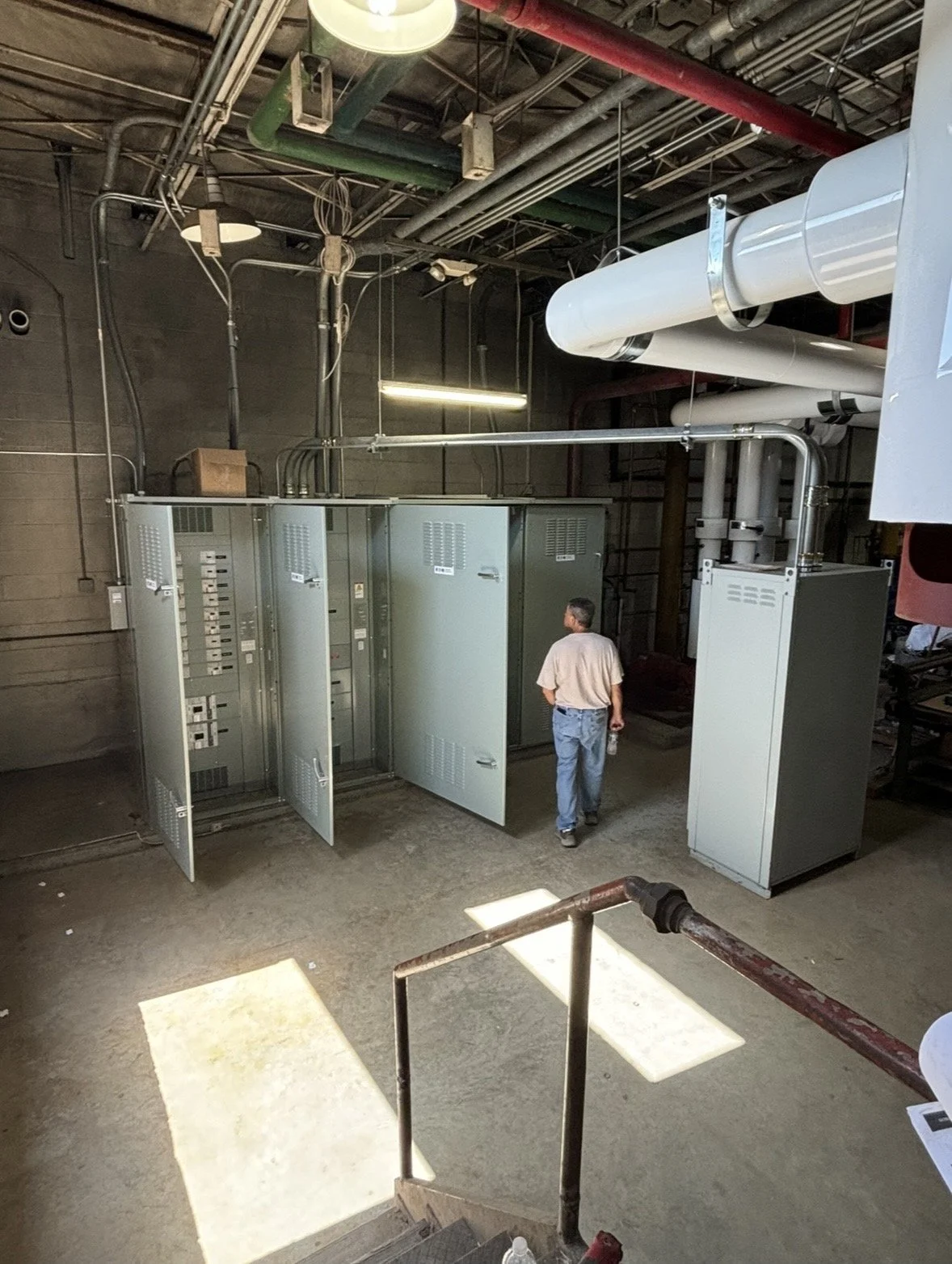 Industrial mechanical room with electrical control cabinets, overhead ventilation ducts and pipes, and a maintenance worker.