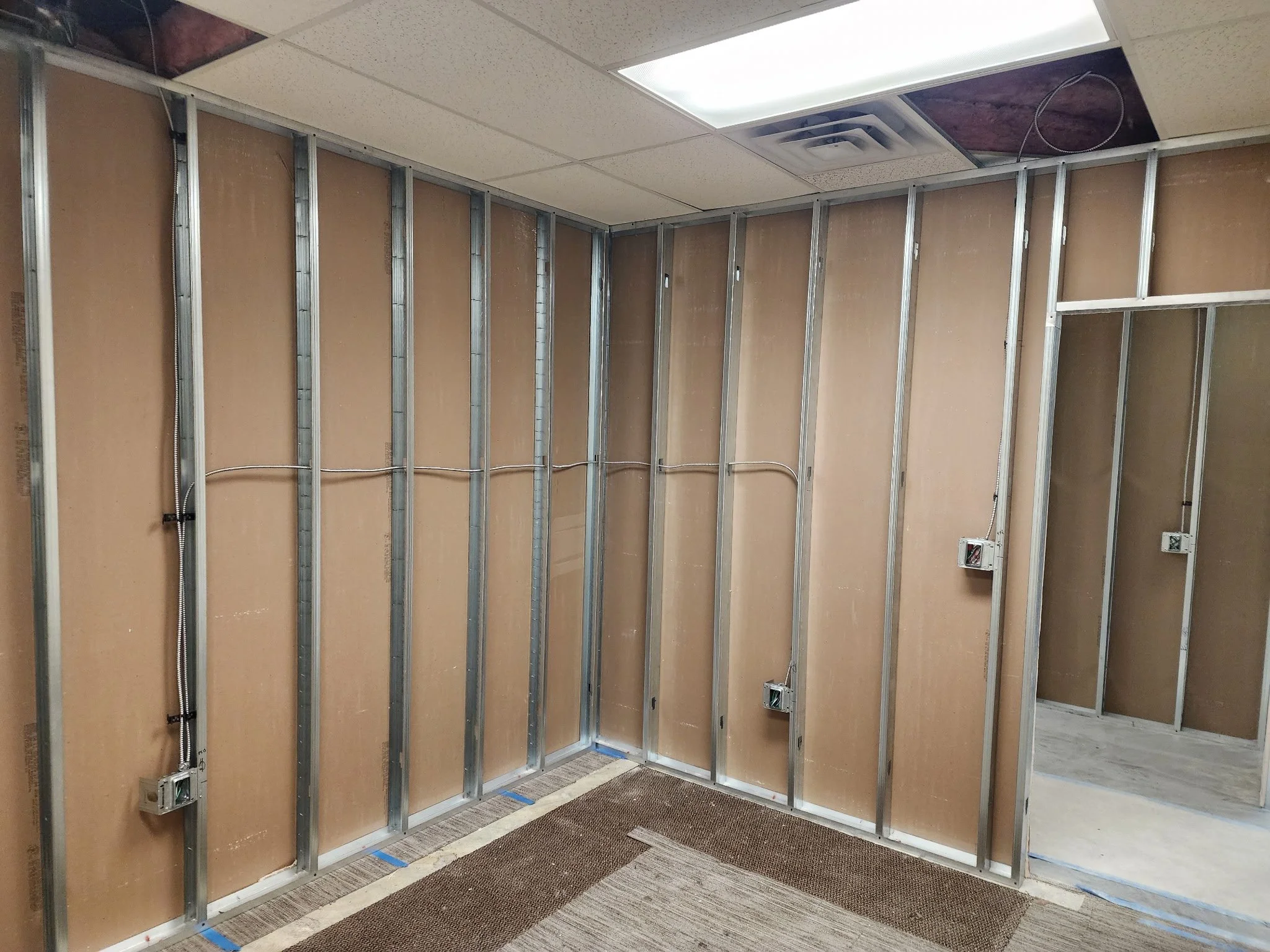Unfinished interior room with exposed metal wall studs, electrical wiring and outlets, partially installed drop ceiling, and construction flooring.