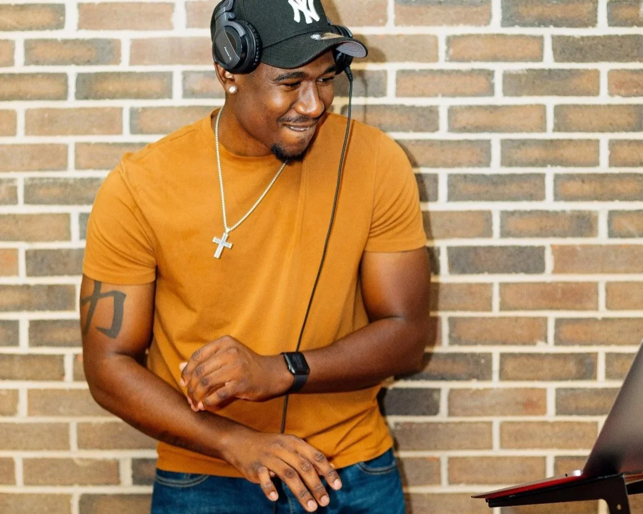 A young man wearing a black cap with a white 'NY' logo, black headphones, a gold chain with a cross pendant, an orange T-shirt, and a tattoo on his right arm, standing in front of a brick wall and smiling while looking at a laptop.