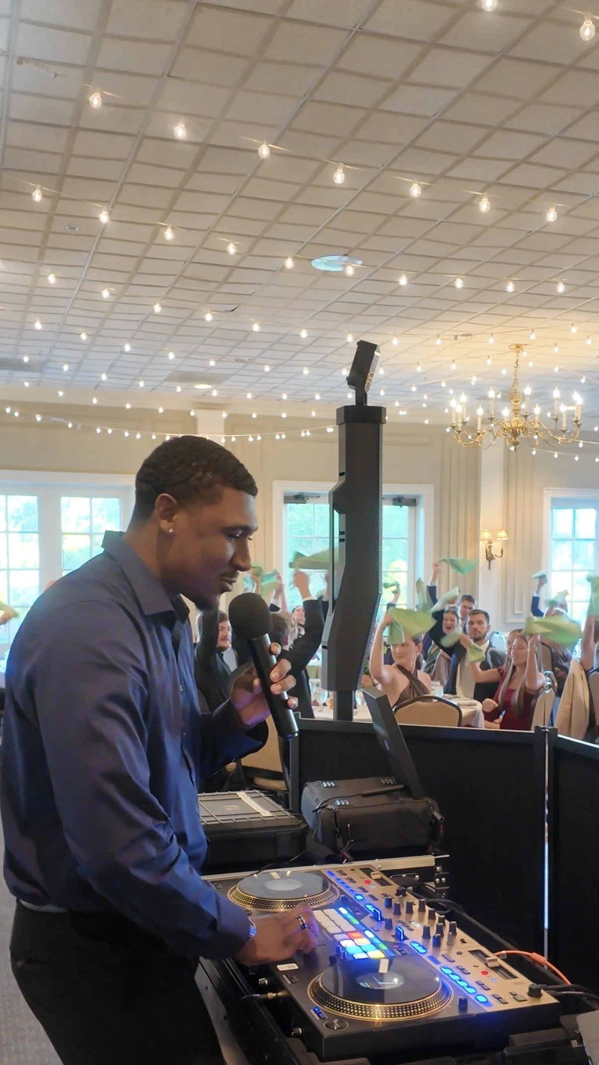 A DJ in a dark shirt plays music with a microphone and DJ equipment at a party or event, with guests in the background holding napkins or cloths, smiling and dancing in a decorated room with string lights and chandeliers.