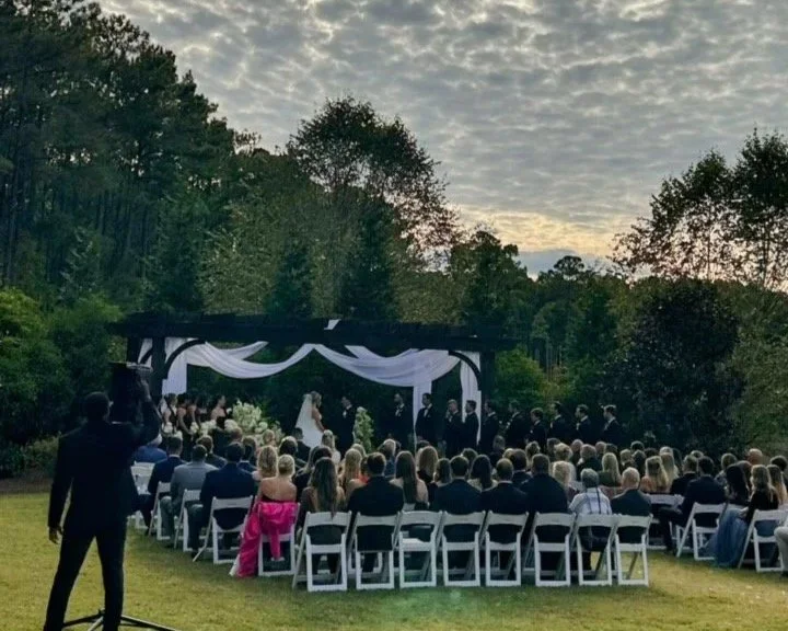 Outdoor wedding ceremony with a large group of guests seated on white chairs, facing a decorated altar with draped white fabric, surrounded by trees during sunset.