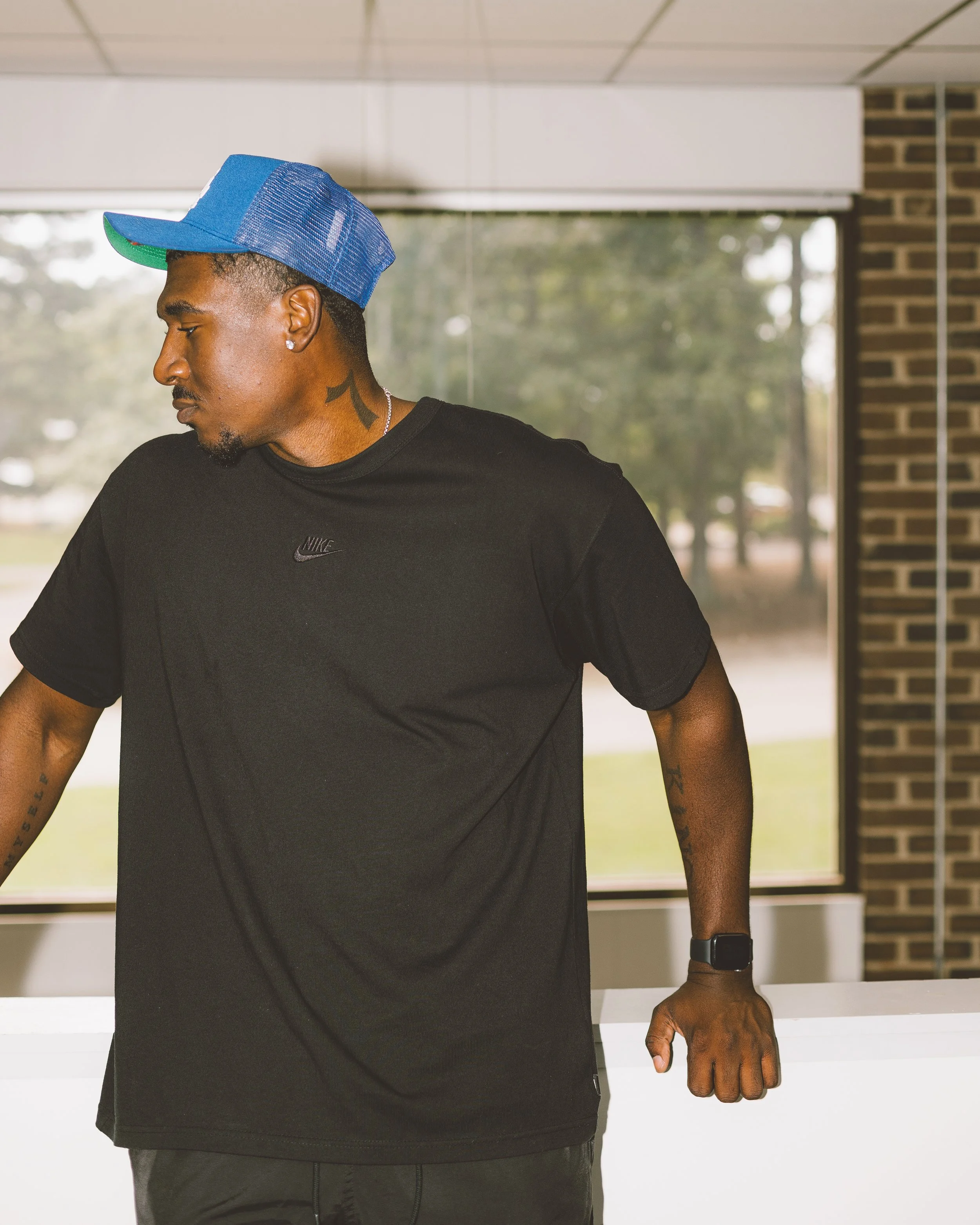 A man wearing a blue cap, black Nike shirt, with tattoos on his neck and arm, and a smartwatch on his wrist, standing indoors with a glass window showing trees outside.
