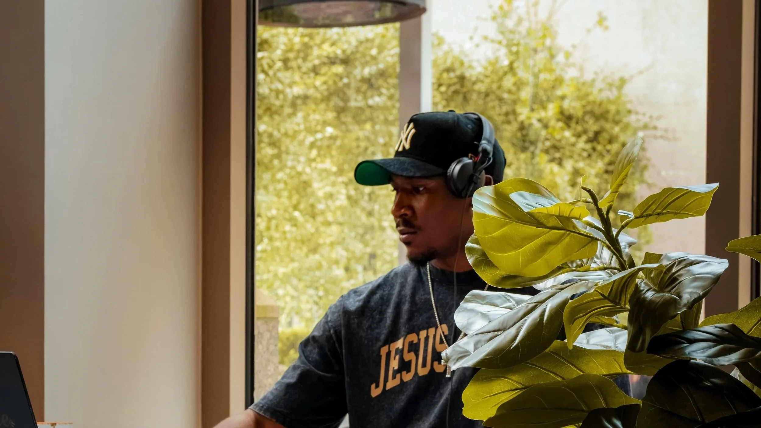 A man sitting indoors near a window, wearing a dark New York Yankees baseball cap, black headphones, and a dark shirt with orange lettering that says 'JESUS'. He appears focused or deep in thought, with a lush green plant partially in the foreground.