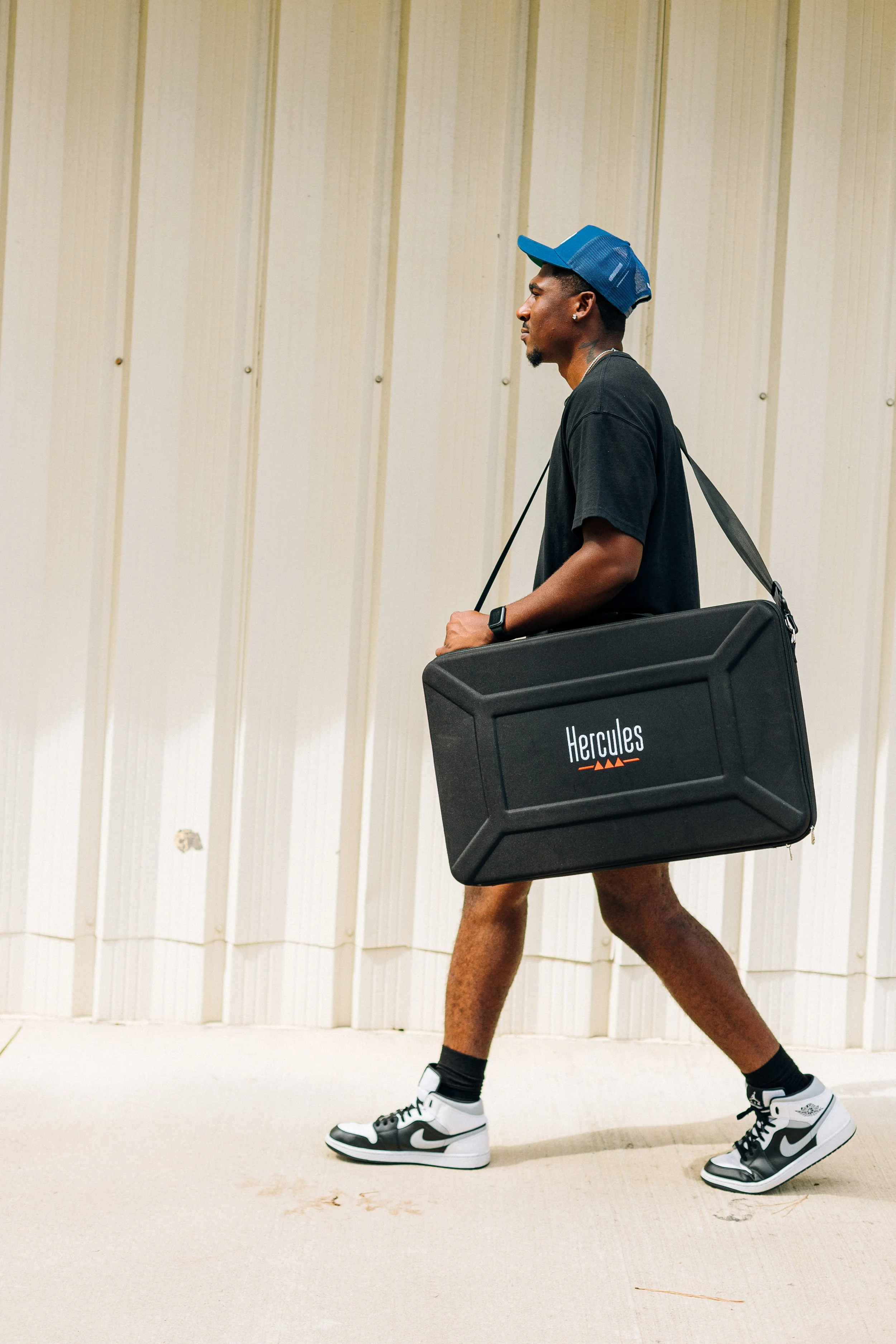 Man walking outdoors carrying a large black case labeled "Hercules." He is wearing a black t-shirt, black and white sneakers, a blue and black cap, and a smartwatch.