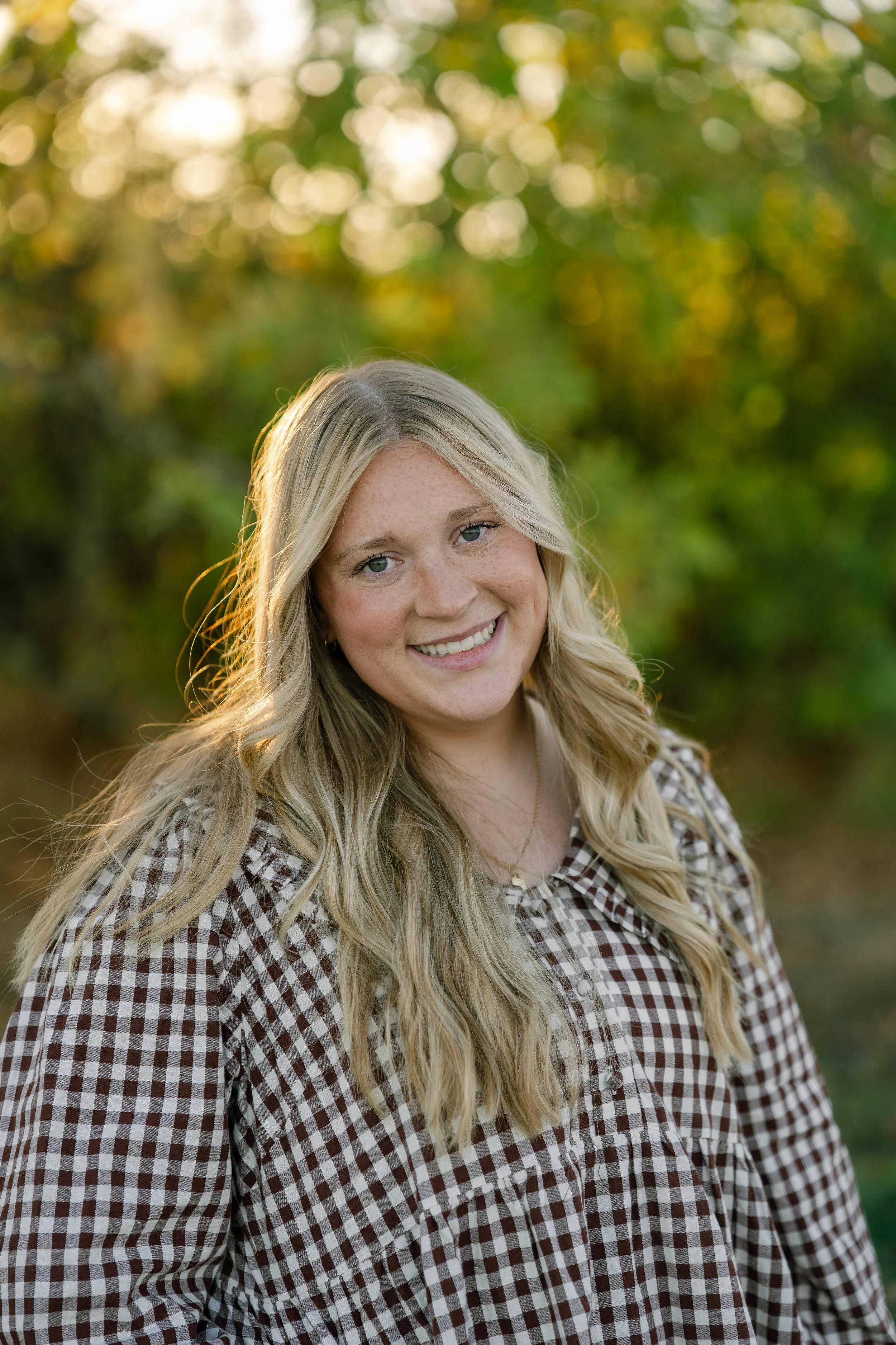 Smiling blonde woman in a brown-and-white checkered shirt outdoors with a sunlit, blurred green background