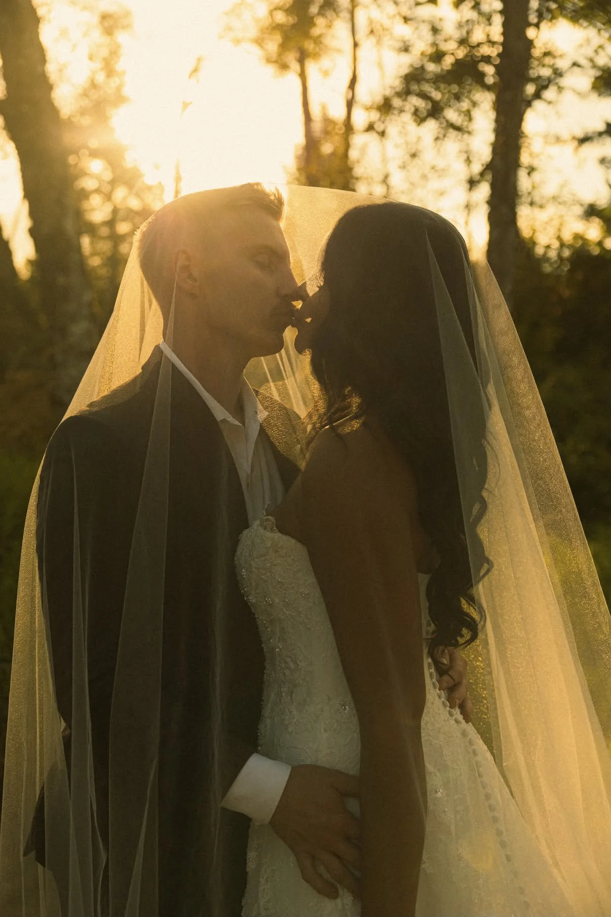 Newlywed couple kissing beneath a wedding veil in a sunlit Oregon forest.