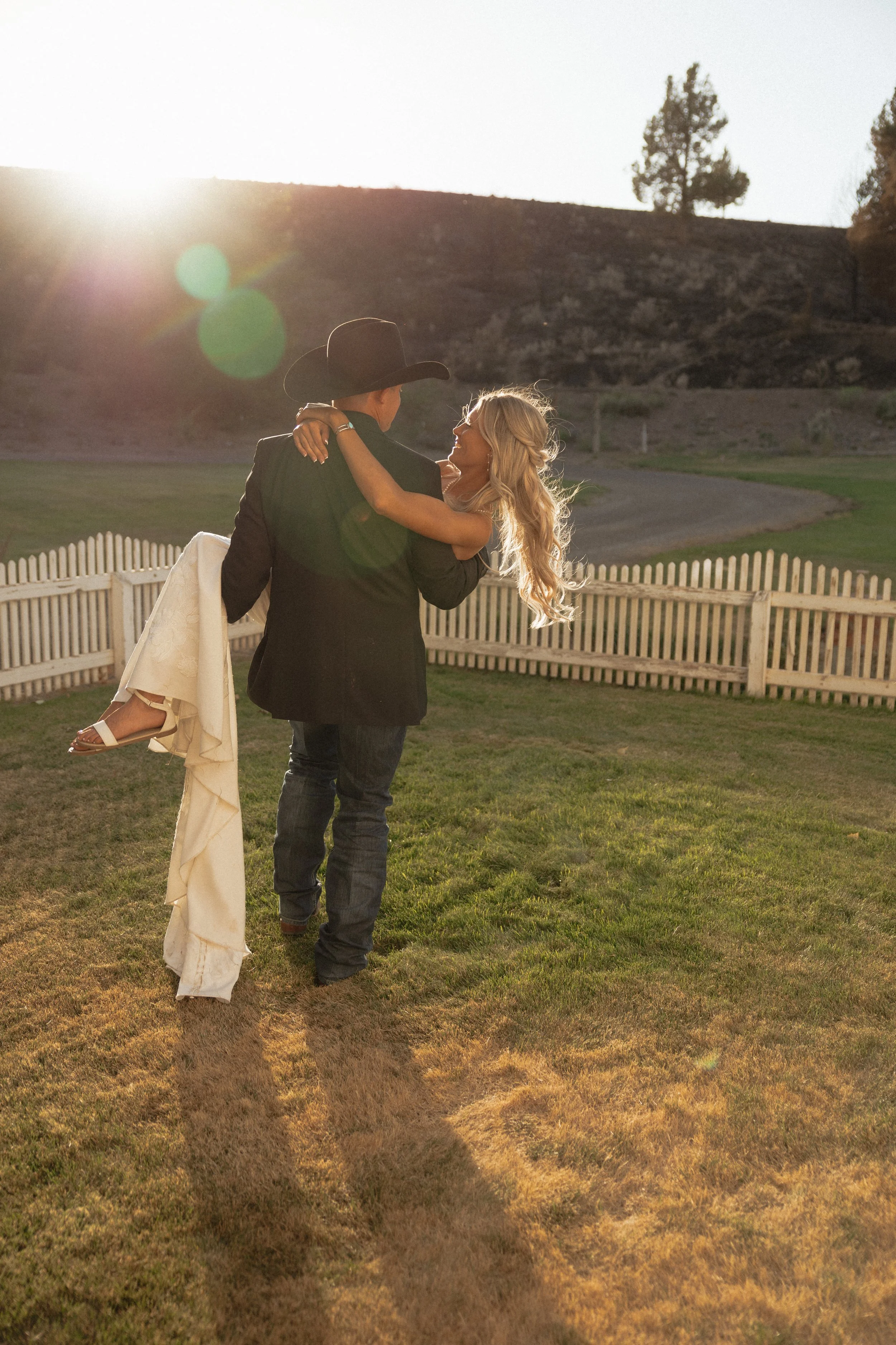 Groom in a cowboy hat carrying his bride across a grassy field at sunset in Madras, Oregon