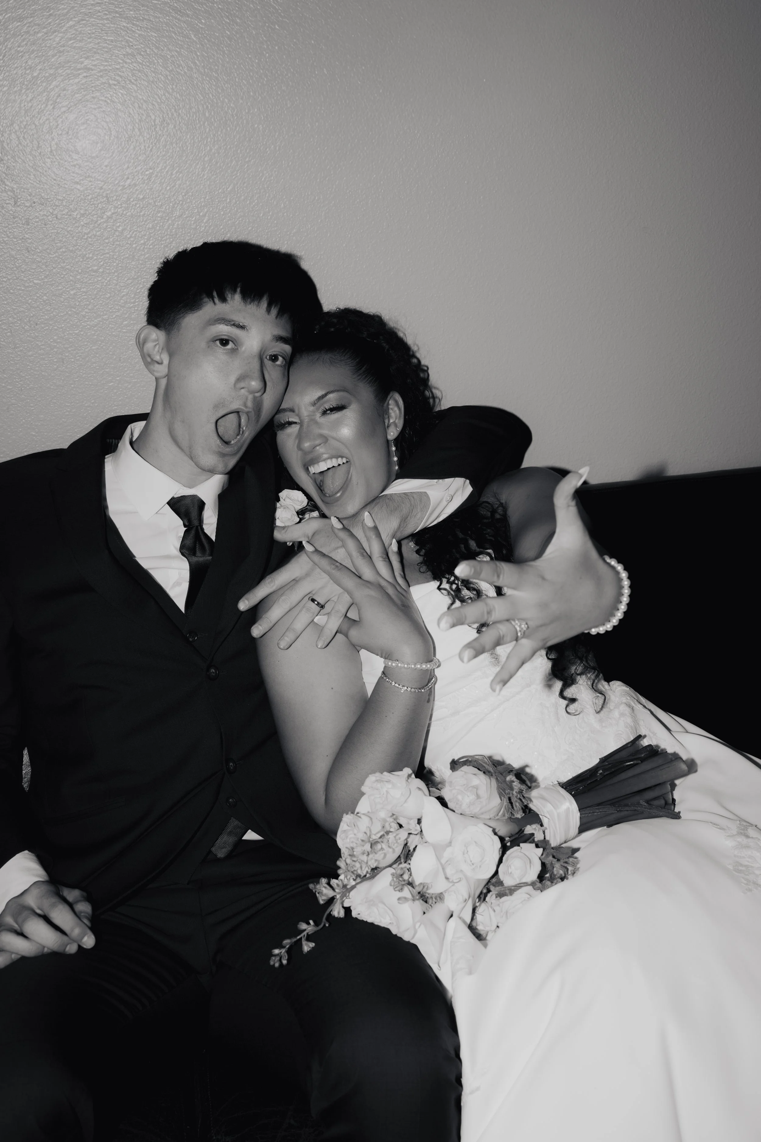 Black-and-white wedding portrait of a laughing bride and groom embracing on a couch with a bouquet of flowers after signing marriage license