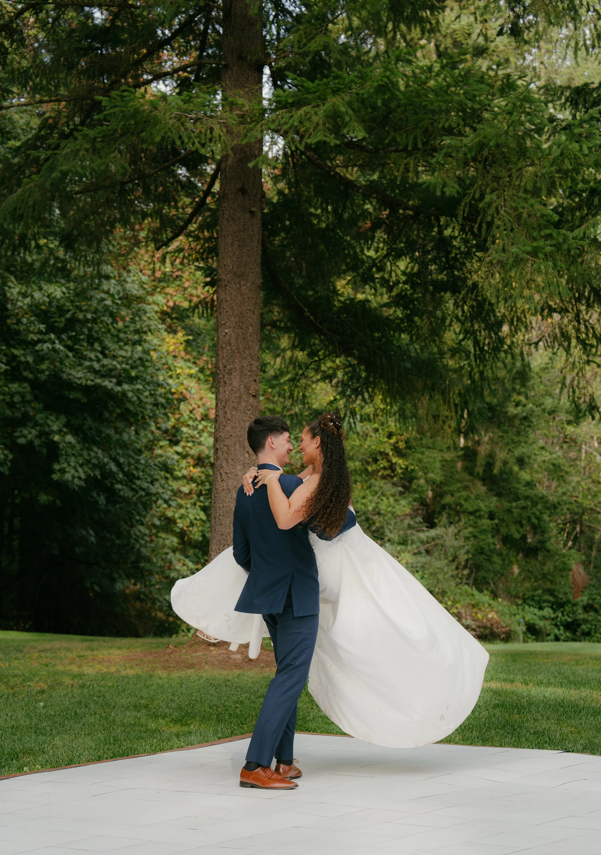 Groom carrying his bride in a flowing white wedding dress outdoors beneath evergreen trees in Oregon for their first dance.