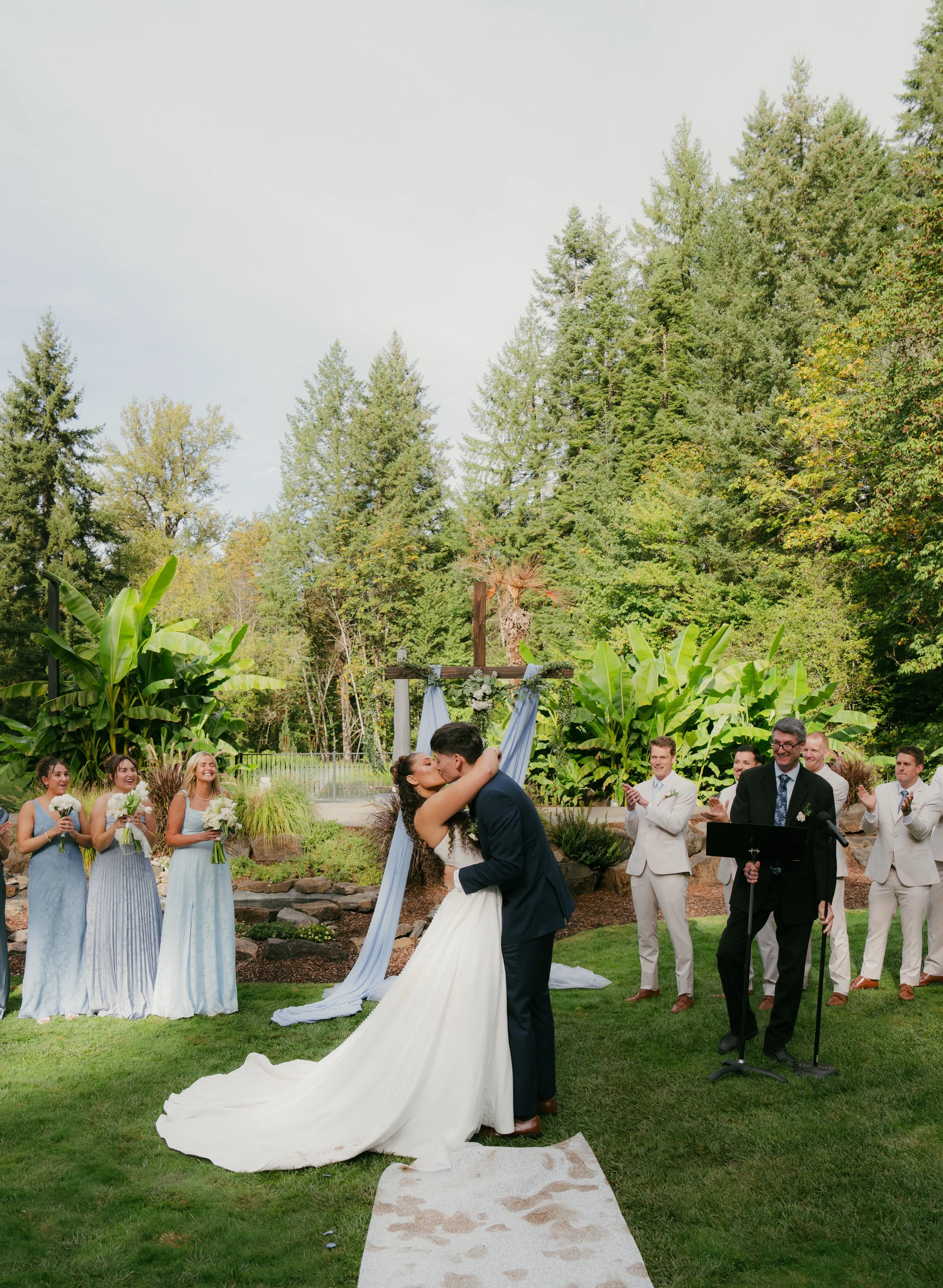 Bride and groom kiss during an outdoor garden in Sweethome, Oregon wedding ceremony as bridesmaids, groomsmen, and guests applaud.