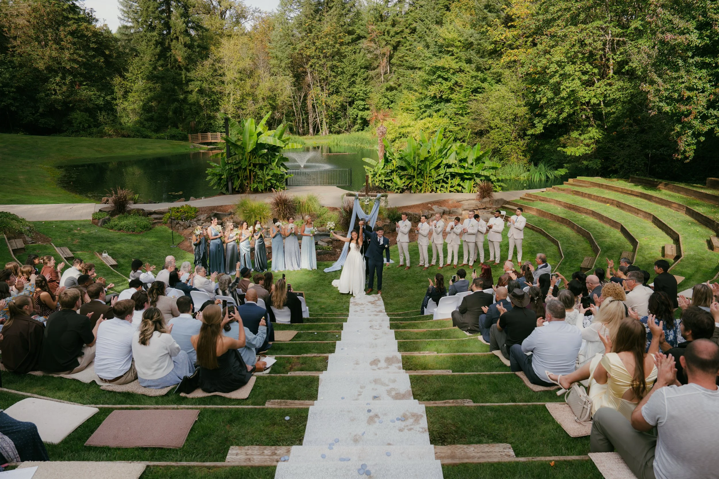 Outdoor wedding ceremony with bride and groom, wedding party, and seated guests beside a landscaped pond and fountain in Sweet Home, Oregon