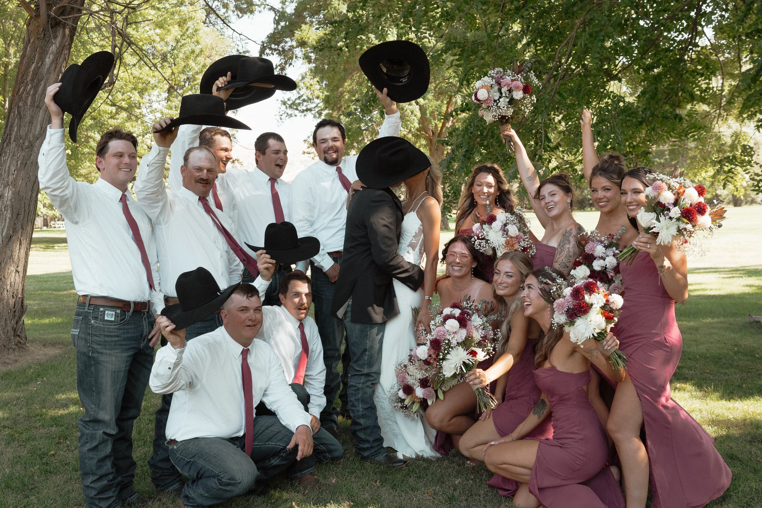 Joyful outdoor wedding party posing under trees, with groomsmen in white shirts and black cowboy hats and bridesmaids holding colorful bouquets in the countryside in Madras, Oregon