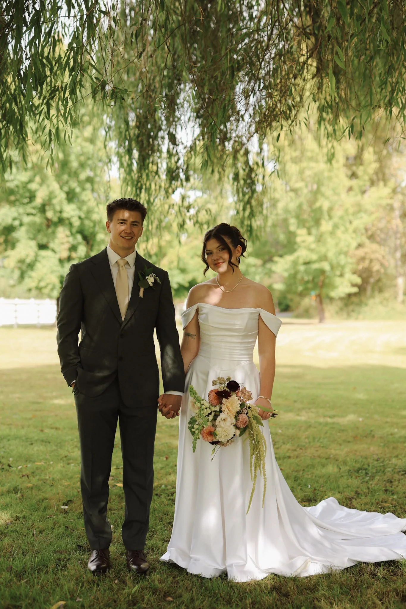 Newlywed couple holding hands outdoors, wearing a black suit and white wedding gown with bouquet