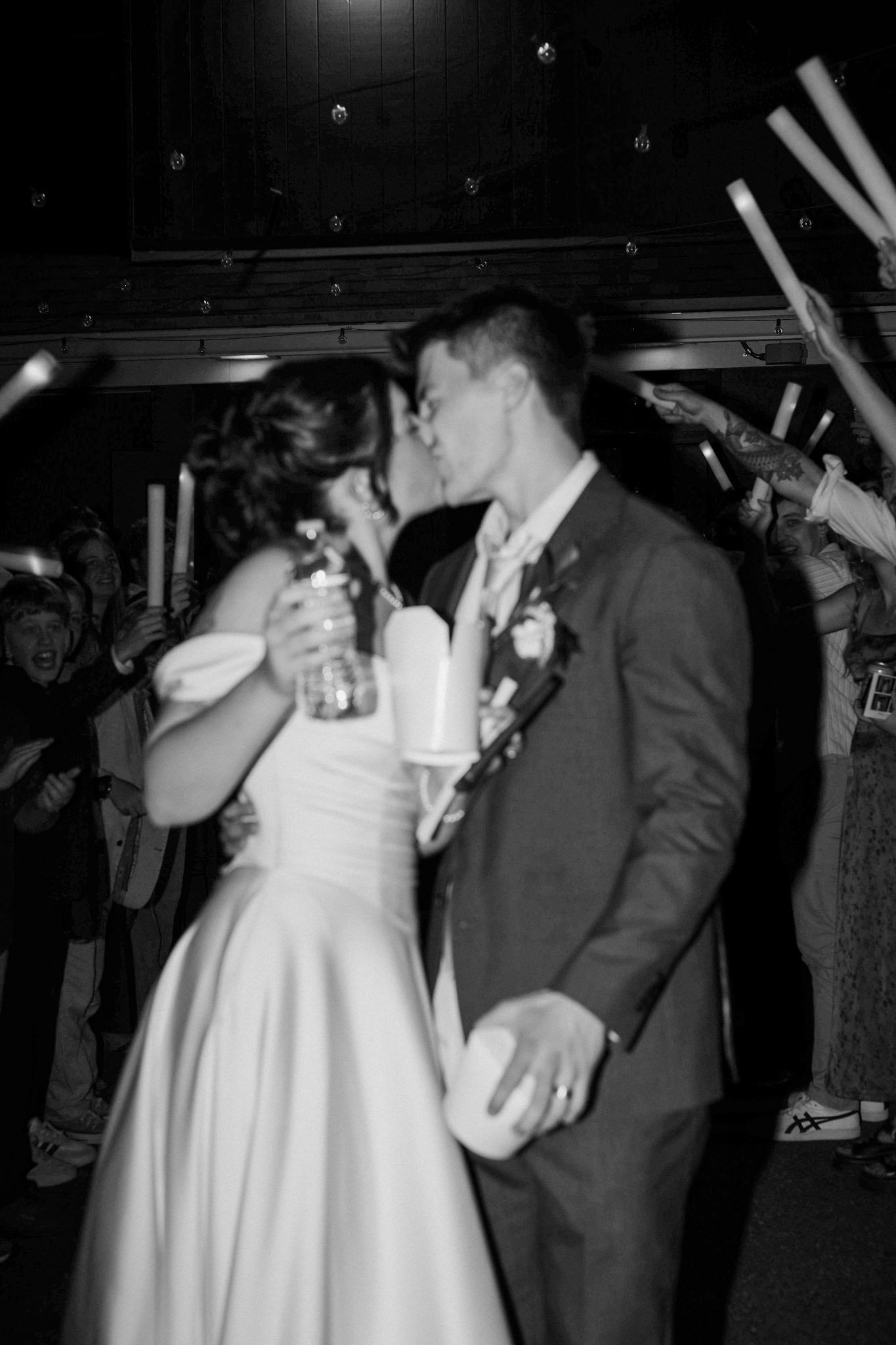 Black-and-white wedding reception photo of a bride and groom are leaving to getaway car while guests send them off with glowsticks
