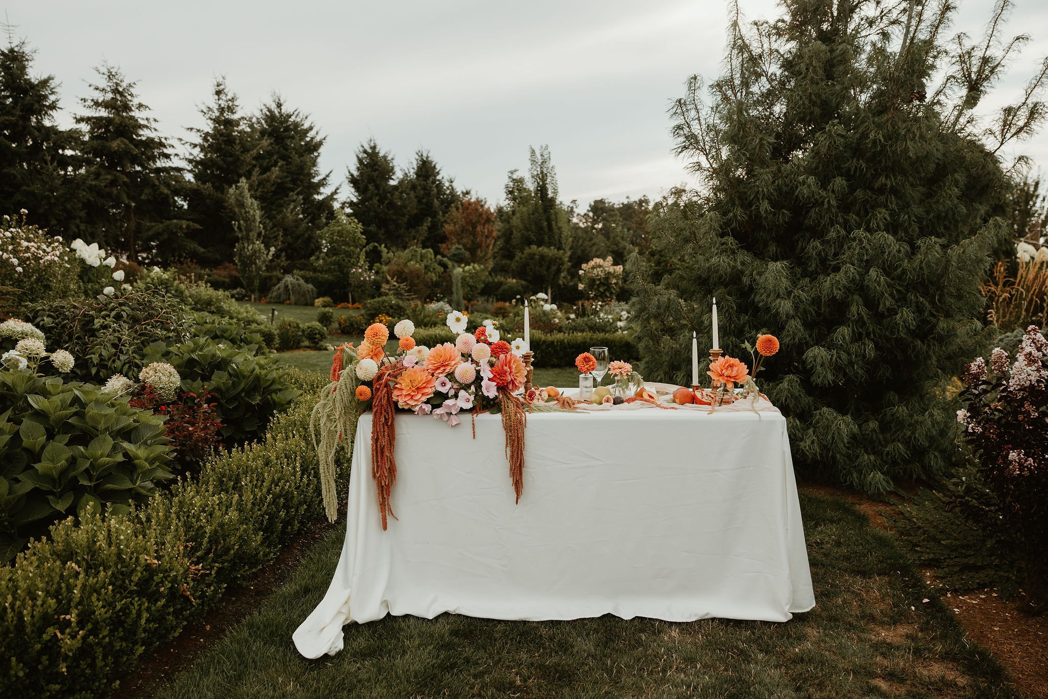 Elegant outdoor wedding reception table with colorful floral centerpiece, white candles, and elegant place settings in a lush garden.