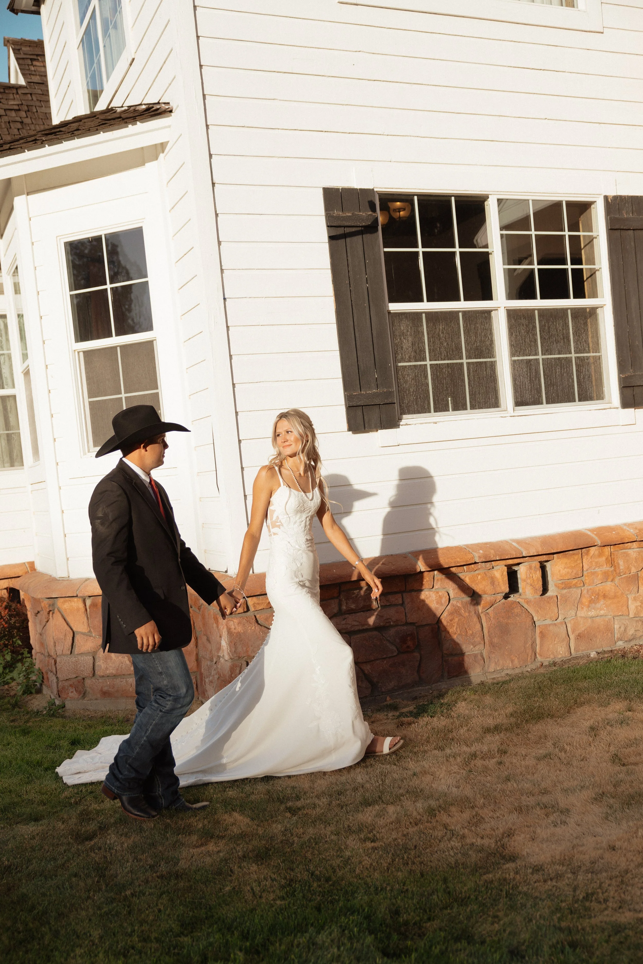 Bride in a white wedding dress and groom in a black cowboy hat walking outside a white house in Oregon