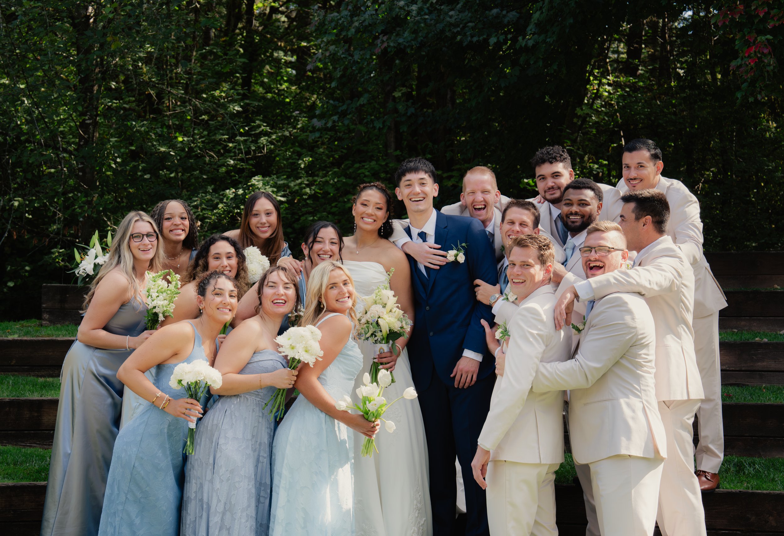 Happy wedding party posing outdoors with the bride, groom, bridesmaids, groomsmen, and white flower bouquets