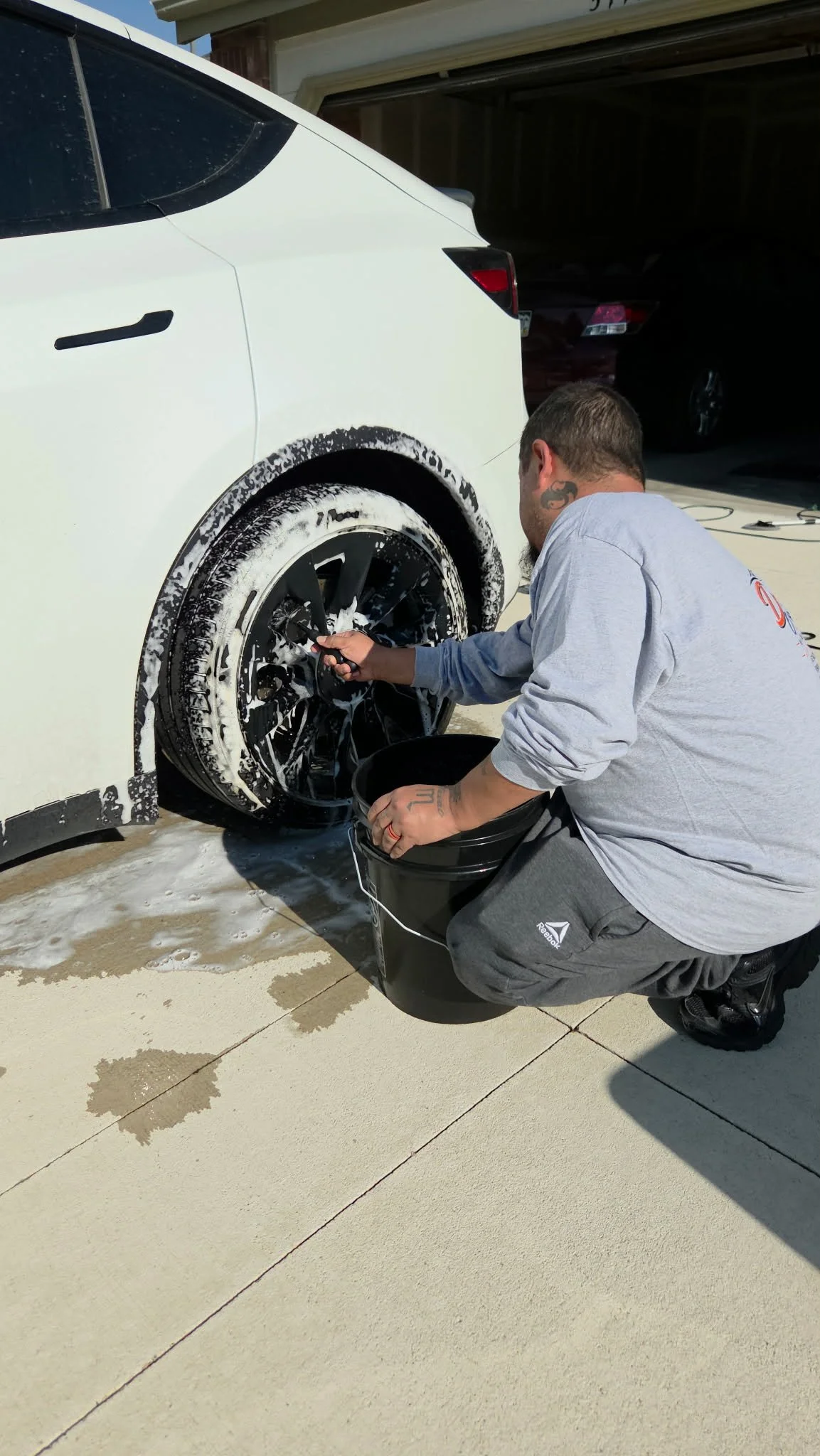 Man washing a white SUV's wheel with soapy water beside a garage.