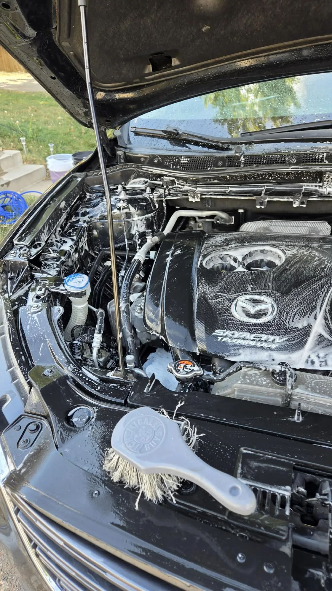 Open Mazda engine bay being washed with soapy water and a detailing brush.