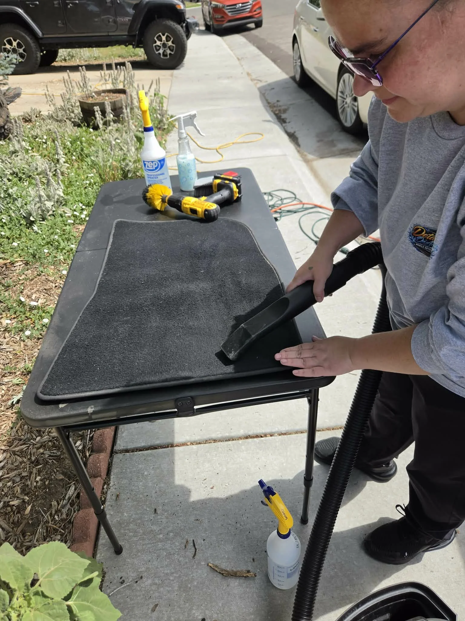 Professional cleaning a vehicle floor mat with a vacuum hose on an outdoor detailing table