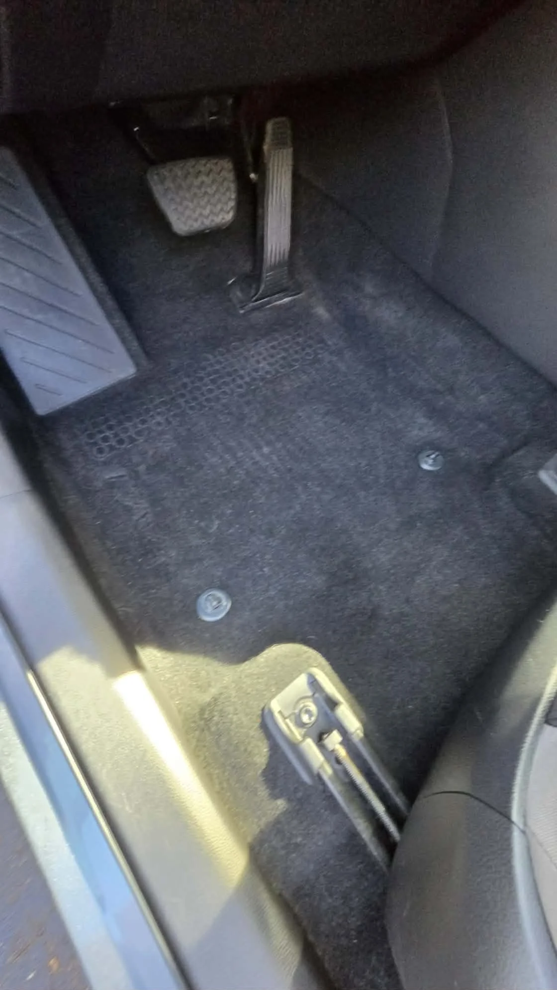 Driver-side footwell of a car showing the brake and accelerator pedals, rubber floor mat, and parking brake lever.