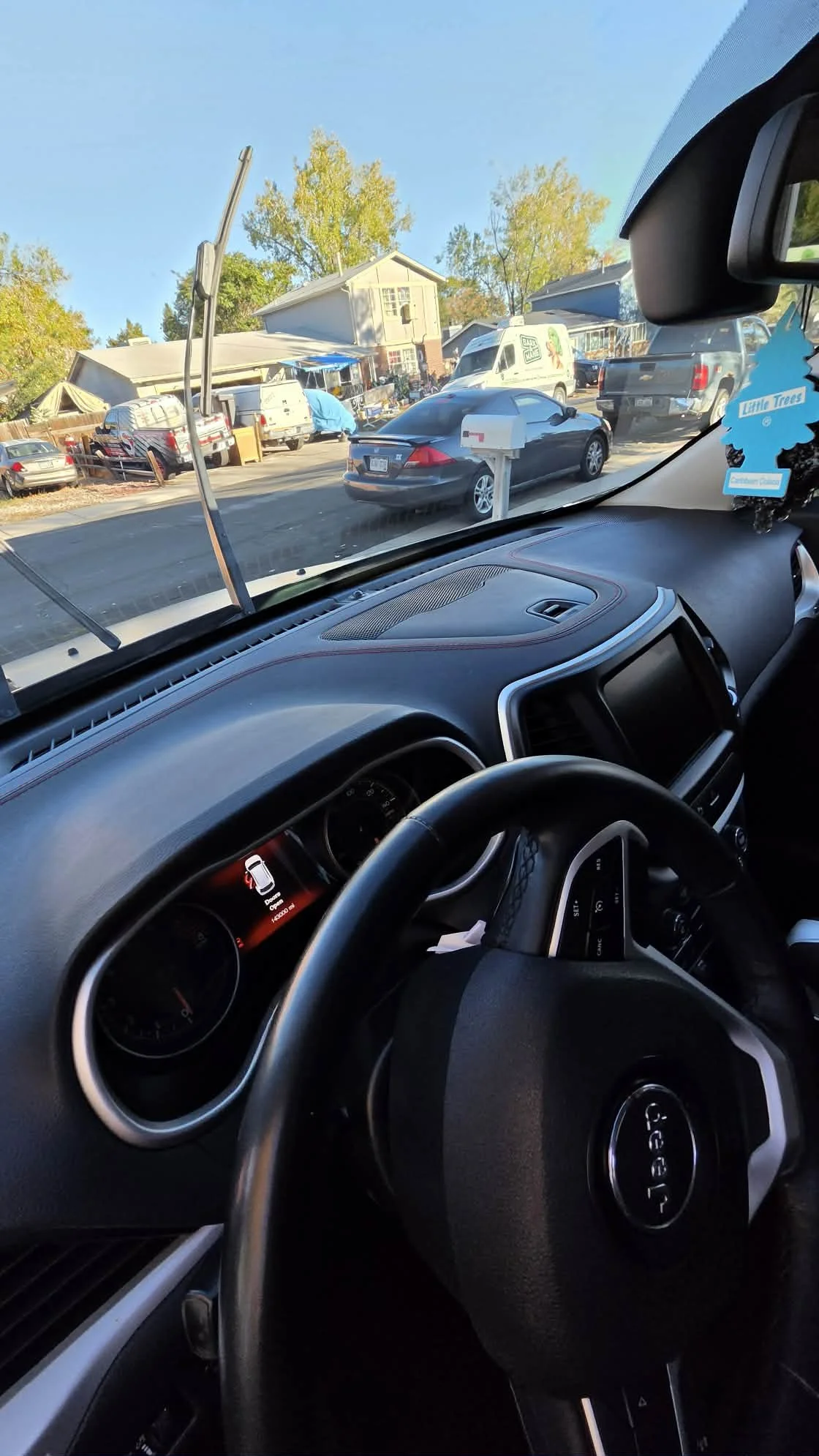 View from inside a Ford vehicle showing the dashboard, steering wheel, Little Trees air freshener, and parked cars outside a suburban home.