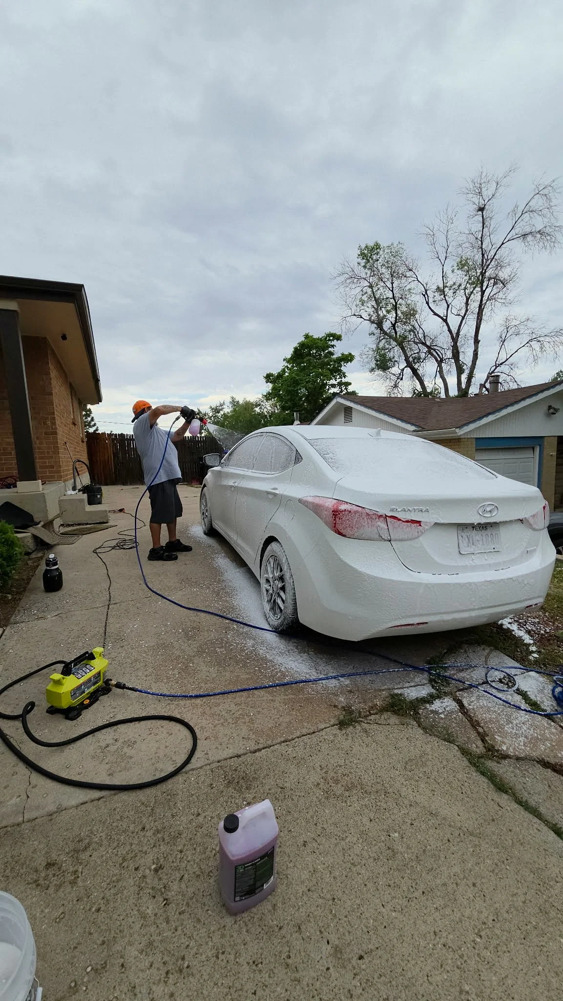 Man pressure-washing a foam-covered white Hyundai Elantra in a residential driveway