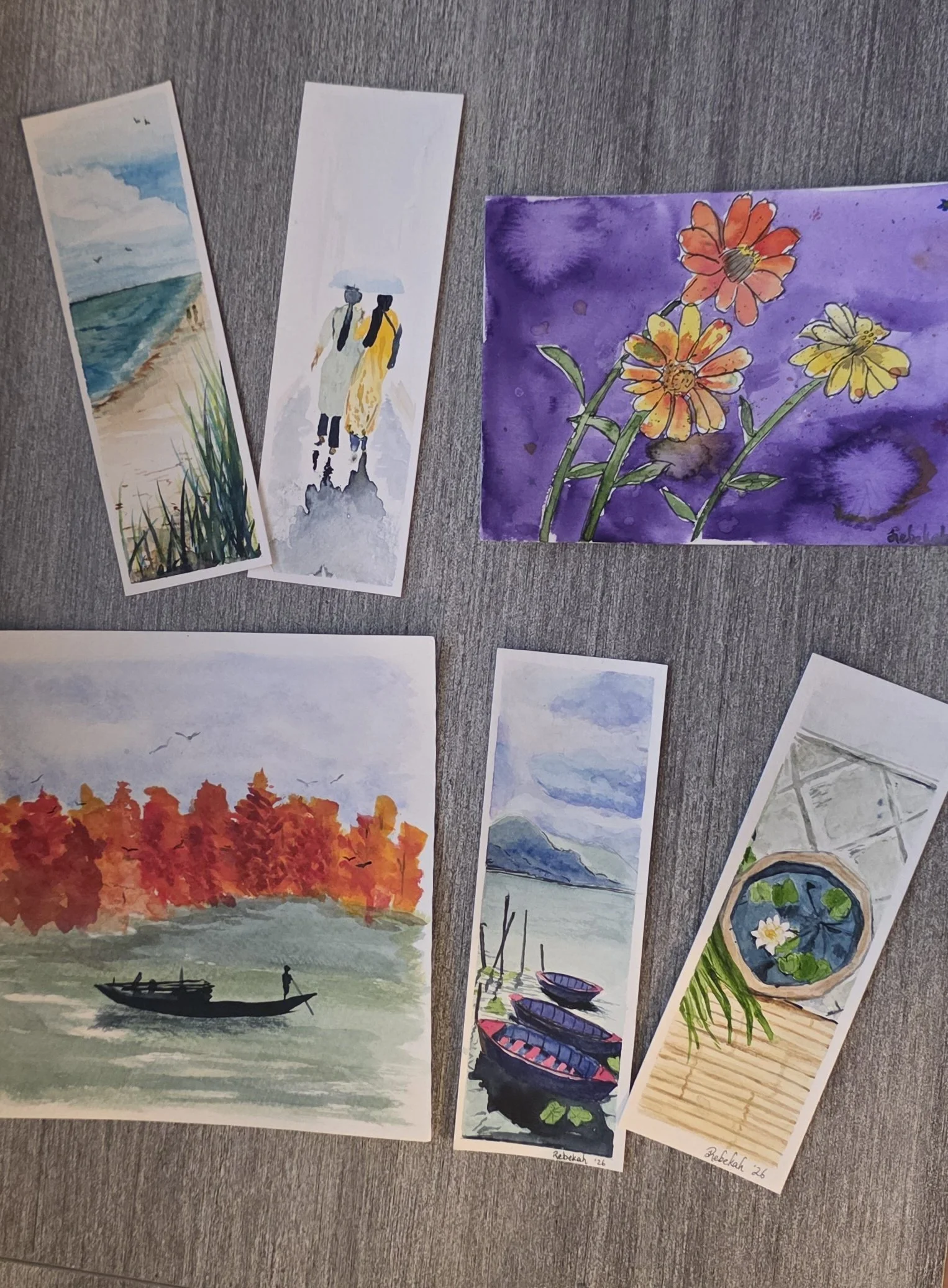 Collection of watercolor paintings on a wood-grain surface, featuring flowers, coastal landscapes, boats, autumn trees, and a pond with water lilies.