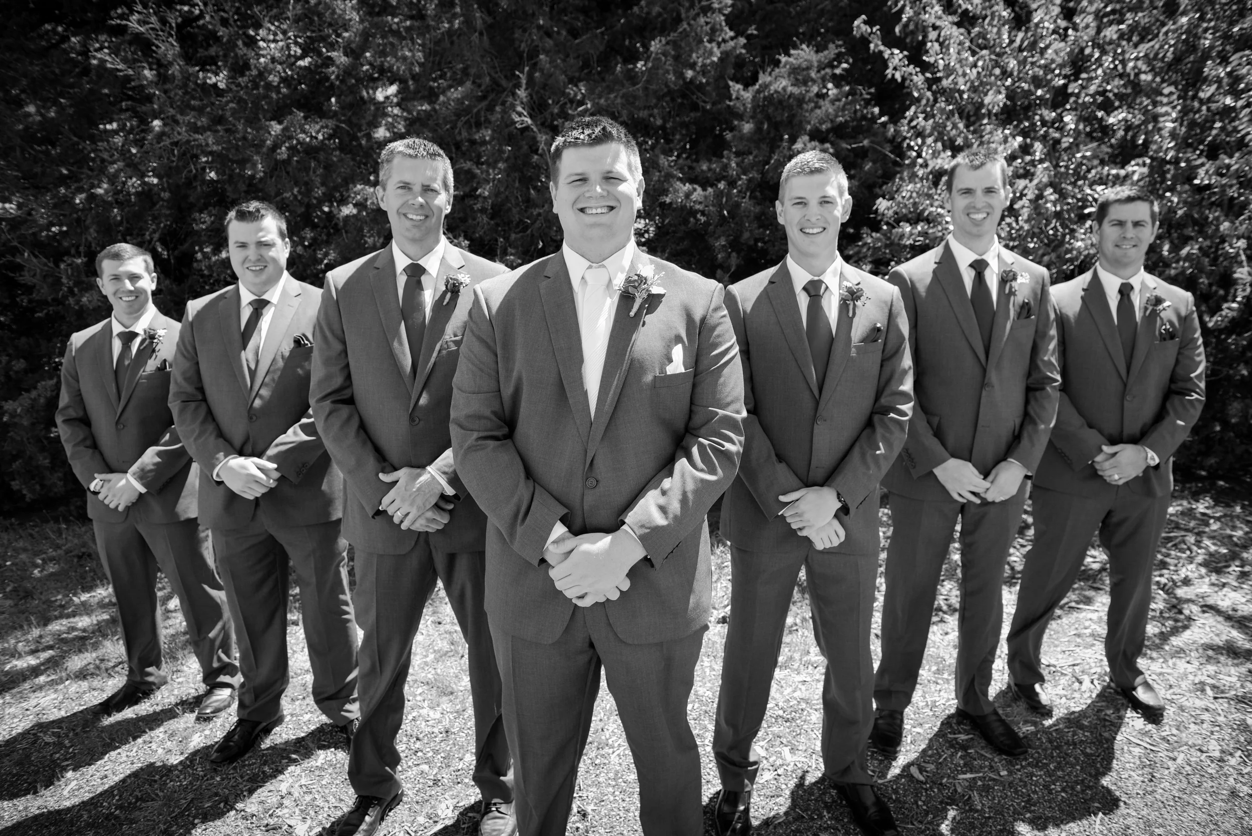 Black and white photo of seven men in suits standing outdoors in front of trees, with the man in the center smiling with hands clasped.