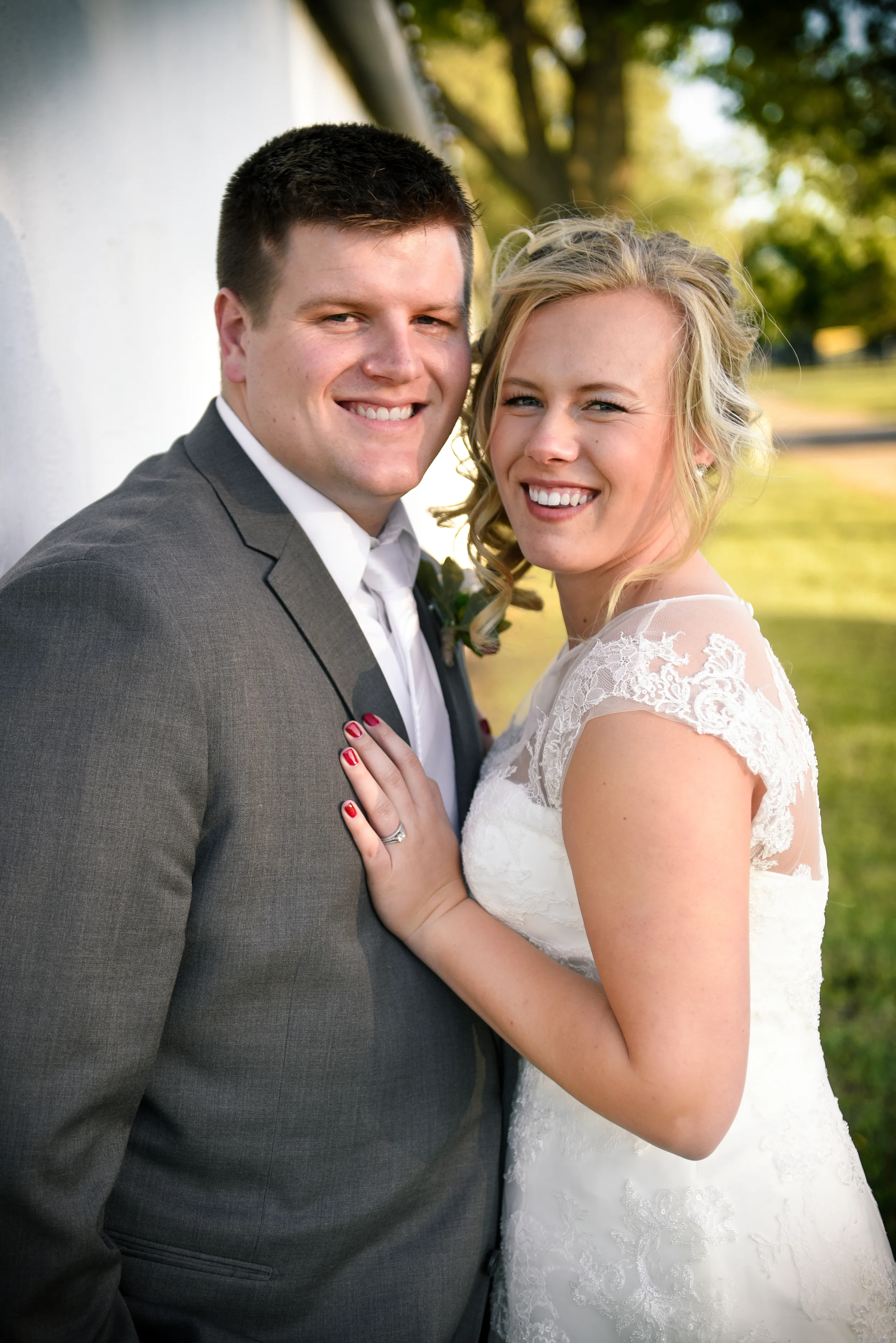 A newlywed couple standing outdoors near a white wall, smiling at the camera. The groom is wearing a gray suit with a white shirt, and the bride is wearing a white lace wedding dress with short sleeves. The bride has blonde hair styled in loose curls