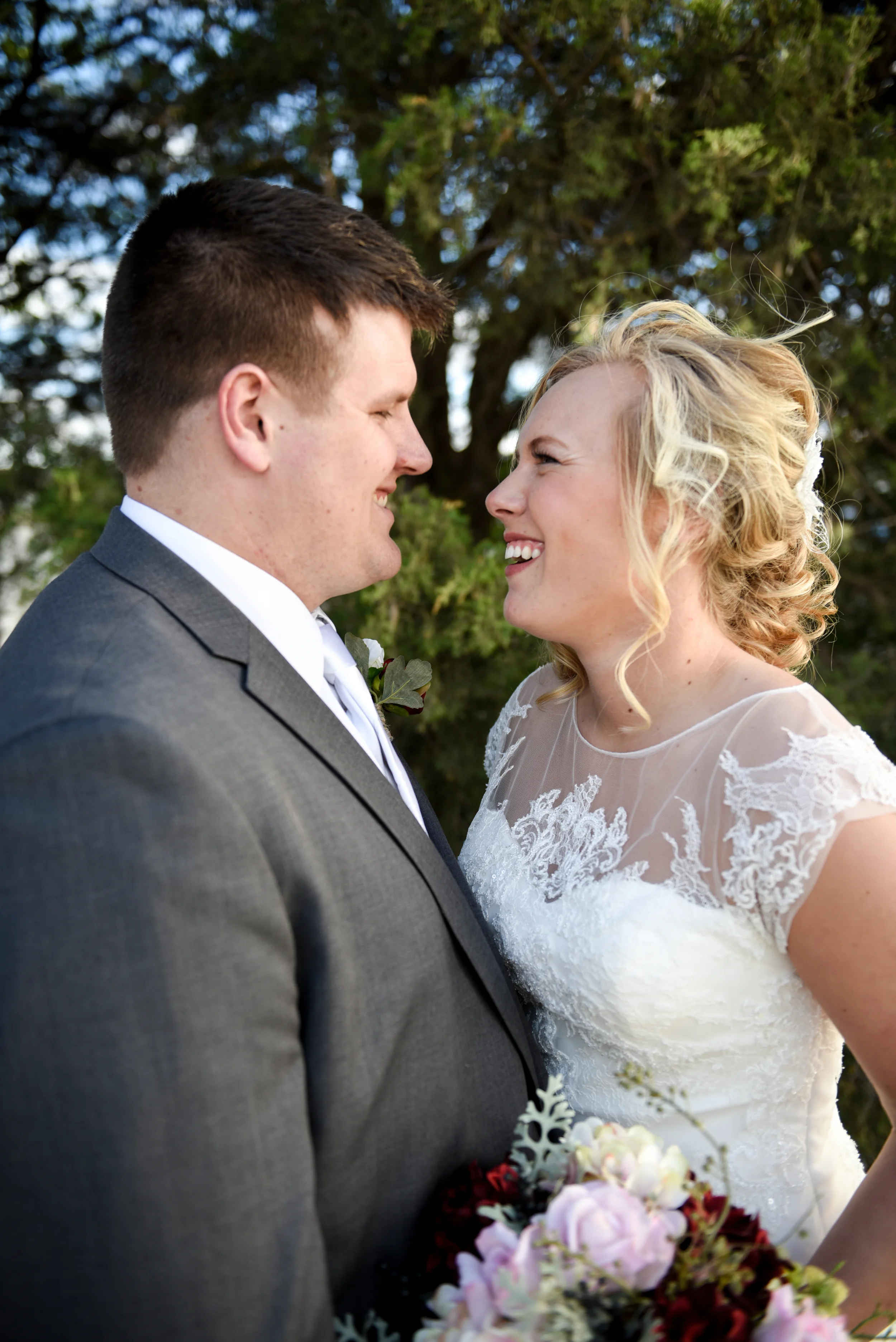 A smiling bride and groom face each other outdoors, with a background of green trees, during a wedding ceremony.