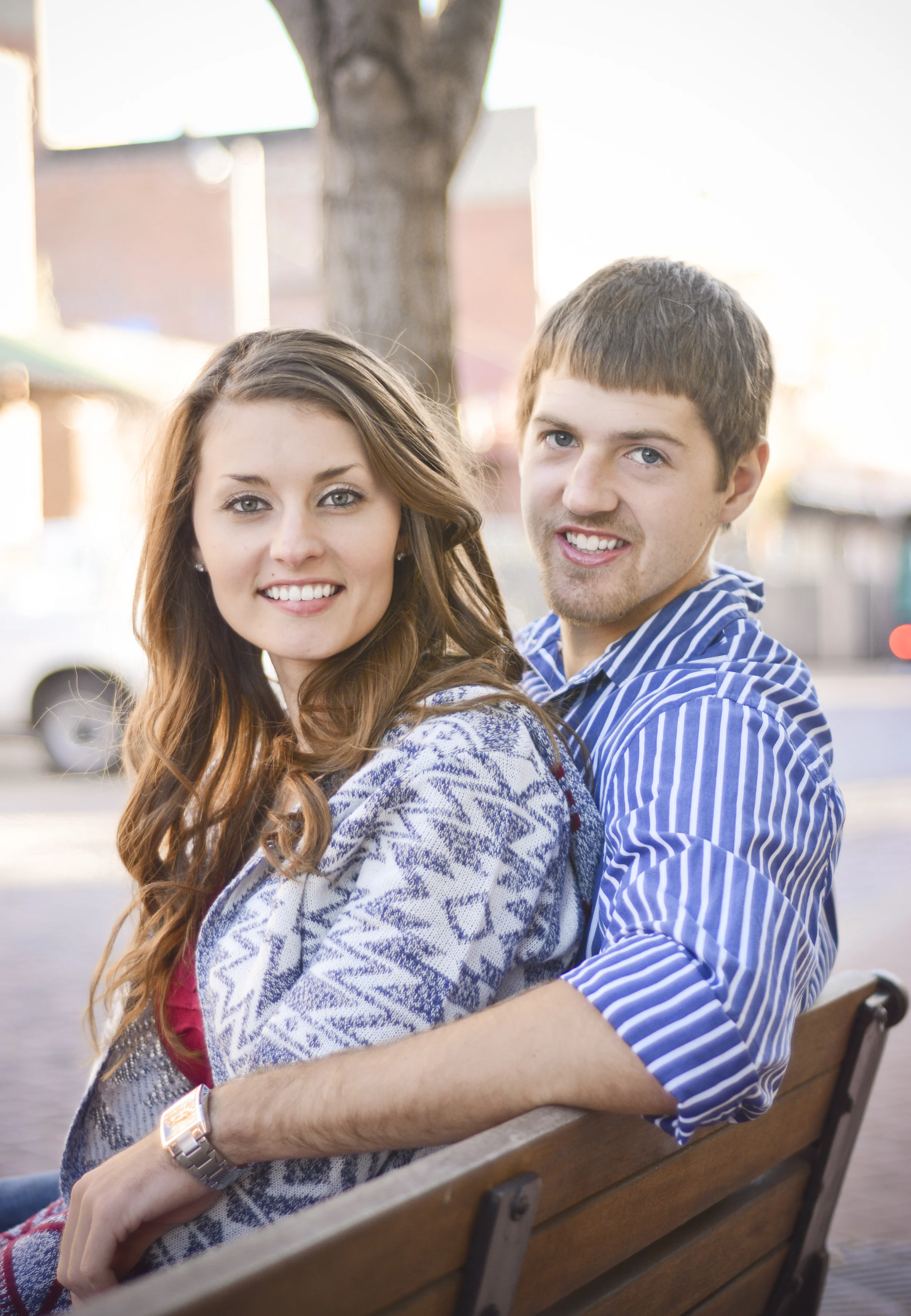 A young couple sitting closely on a park bench, smiling and looking at the camera. The woman has long, wavy brown hair and is wearing a patterned cardigan, while the man has short brown hair and is wearing a striped blue shirt. The background shows a