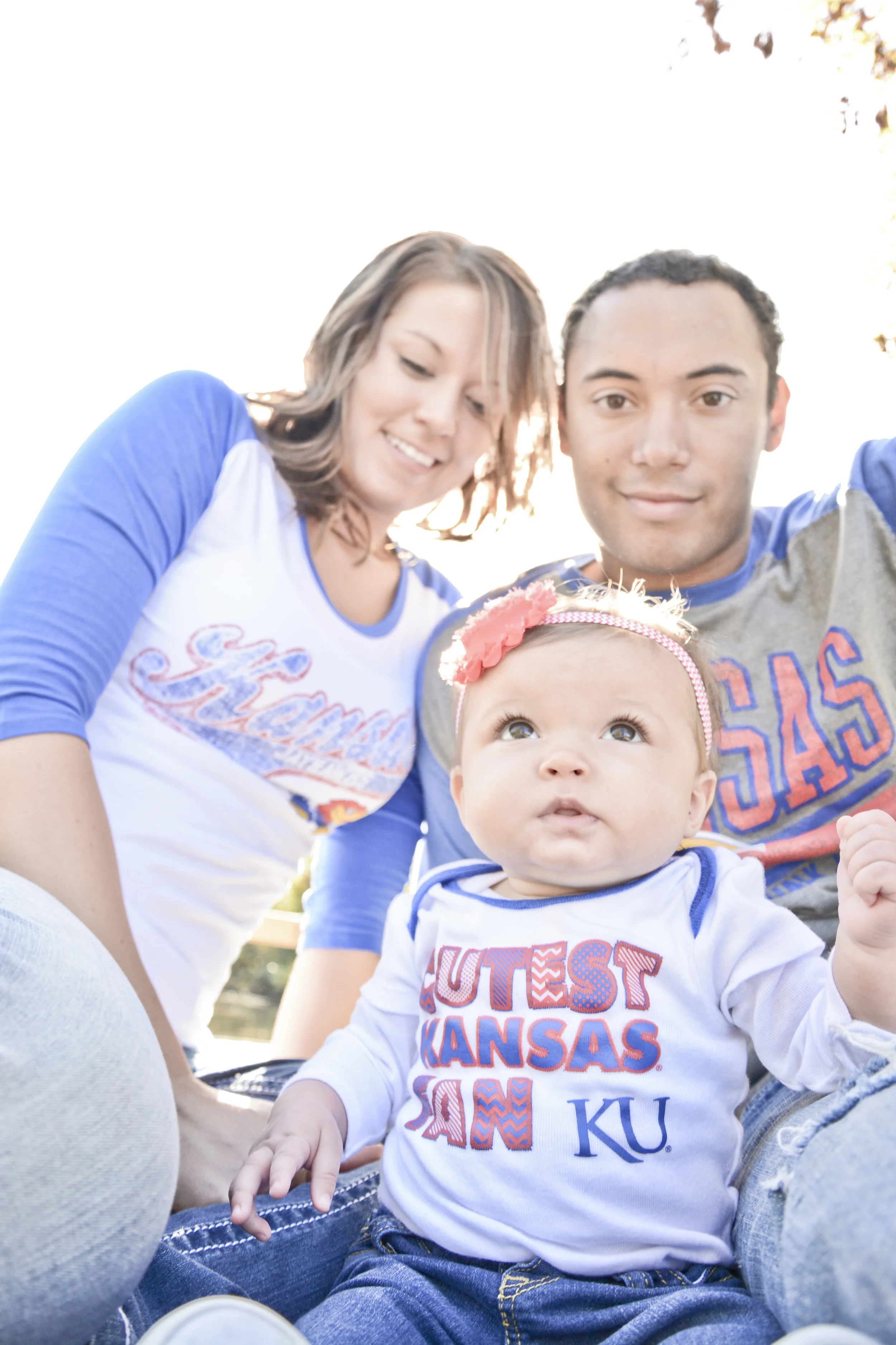 A young family outdoors, sitting together on a sunny day. The mother has shoulder-length brown hair and is wearing a blue and white Kansas shirt. The father has short dark hair and is wearing a gray Kansas T-shirt. Their baby, wearing a white Kansas 