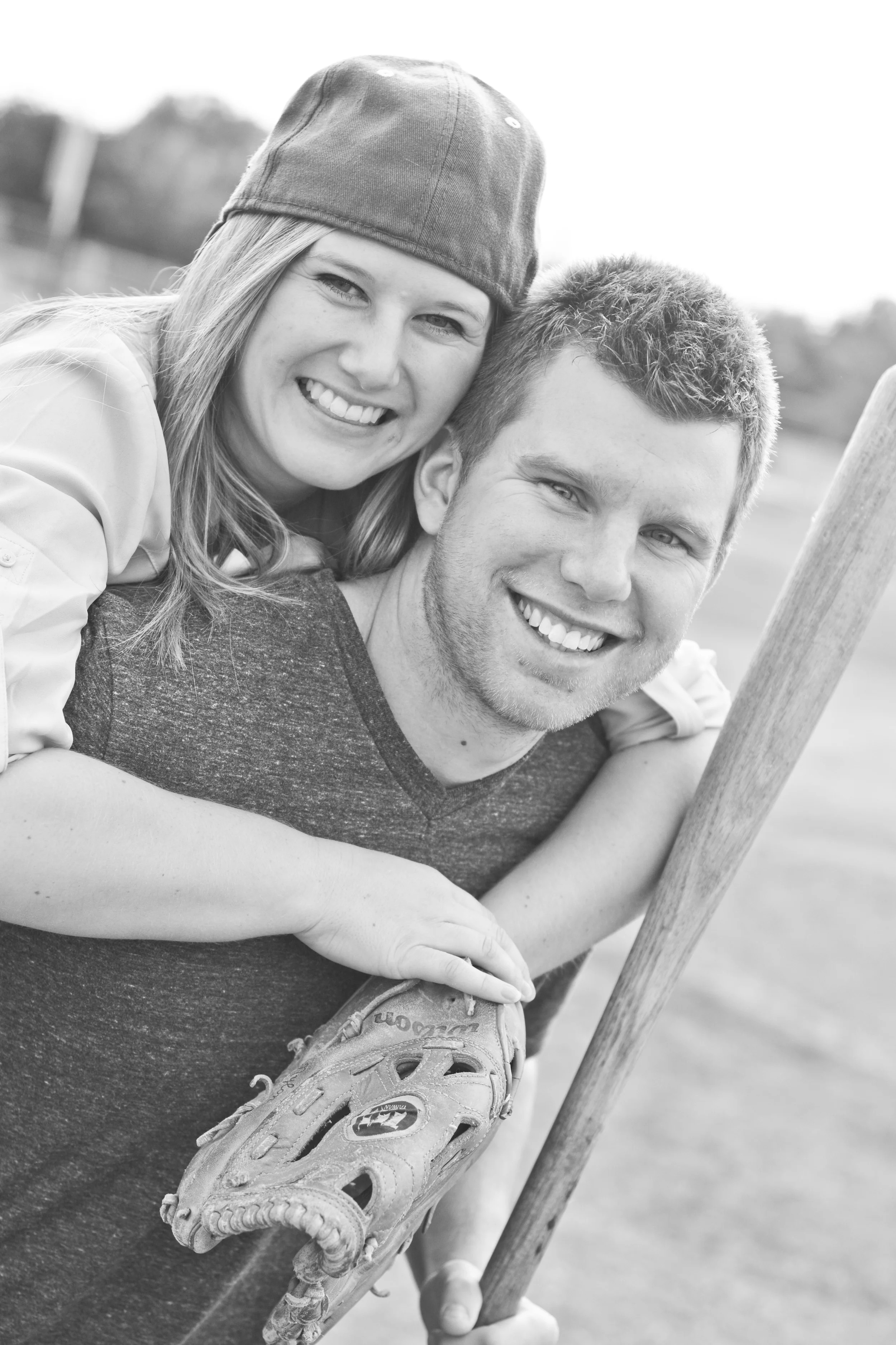 A black-and-white photo of a smiling young woman with long hair, wearing a baseball cap, and a young man with short hair, holding a baseball glove and bat, outdoors.