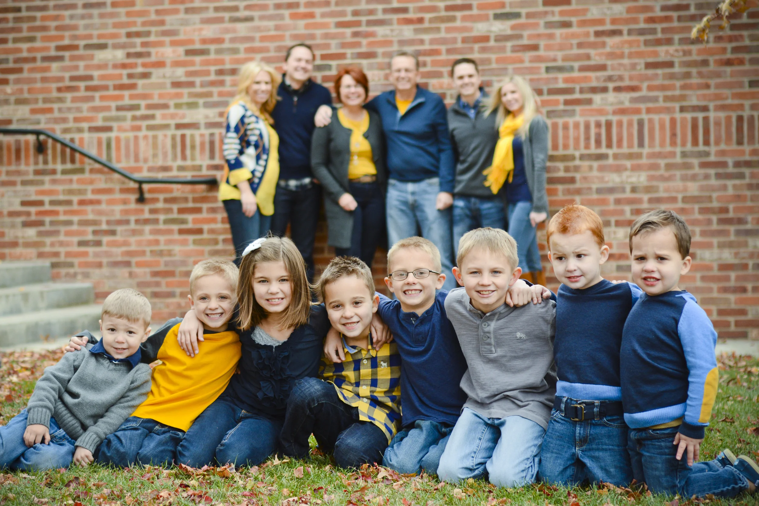 A group of children and adults posing together outdoors in front of a red brick wall, with some sitting on the grass and others standing behind them, during fall.