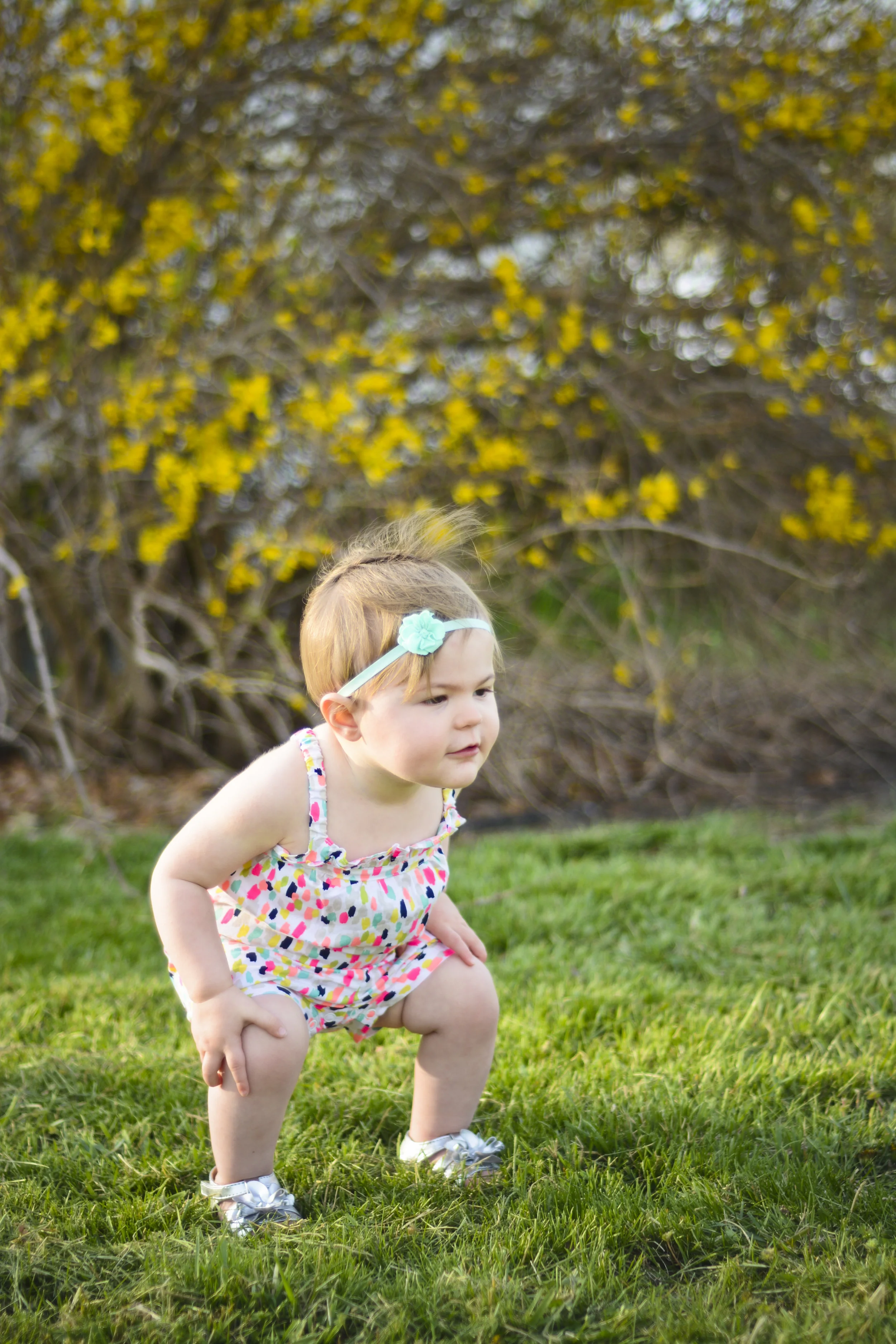 A young girl wearing a colorful dress and silver shoes is squatting on a grassy lawn. She has a headband with a flower and short hair. There are yellow flowers and trees in the background.