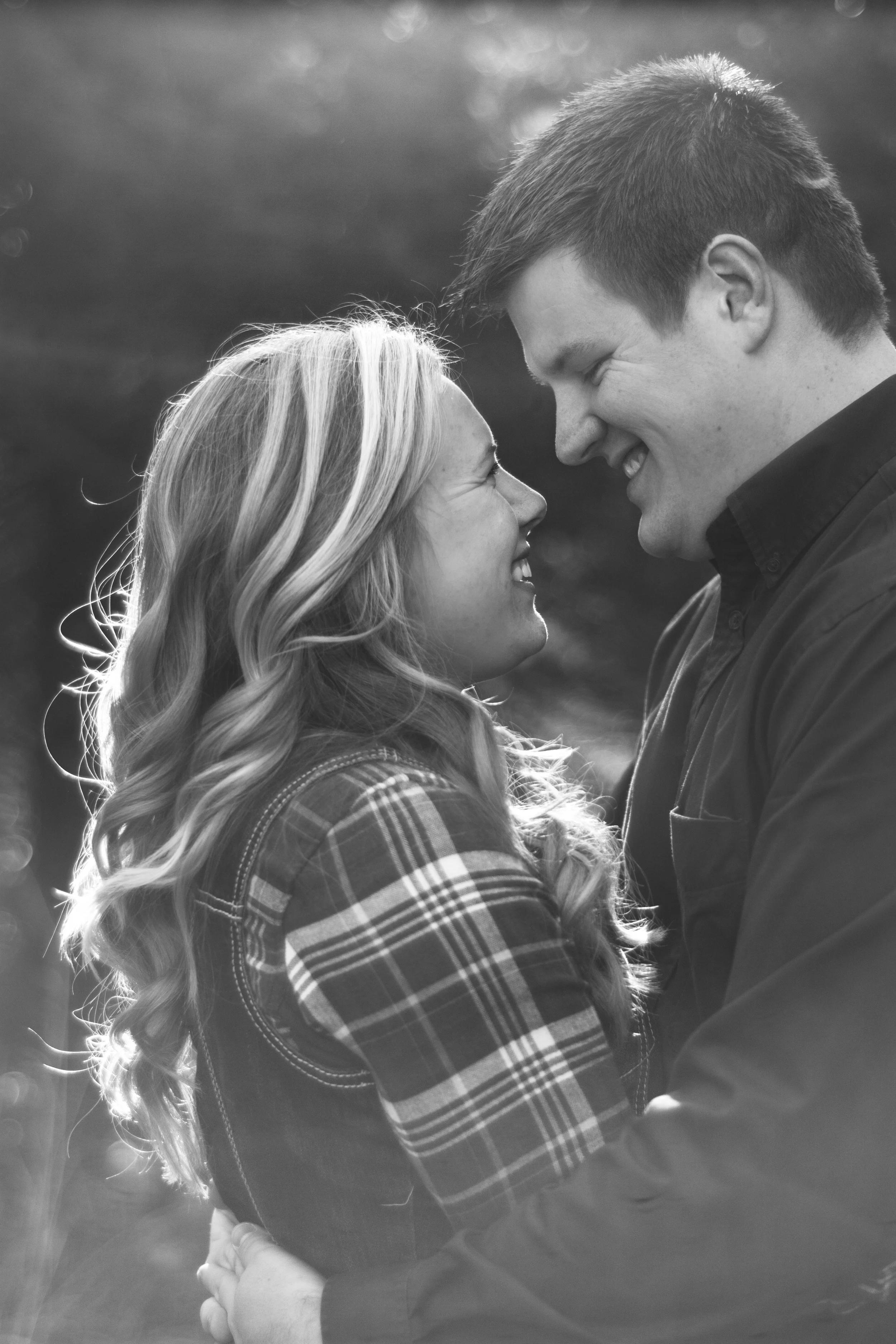 A black and white photo of a couple touching foreheads and smiling at each other outdoors.