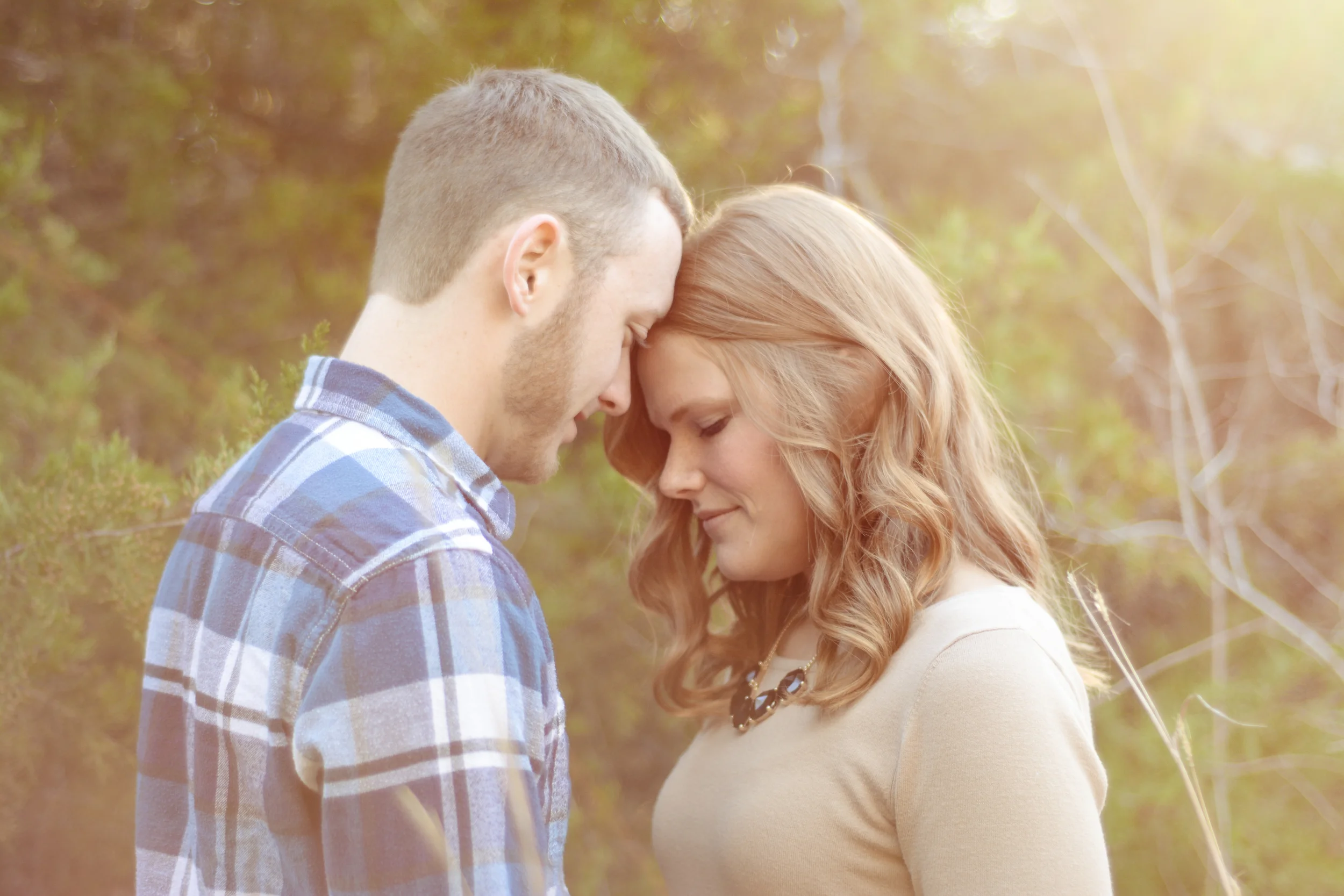 A man and woman standing close with their foreheads touching outdoors in a natural setting, with sunlight shining through trees and blurred foliage in the background.