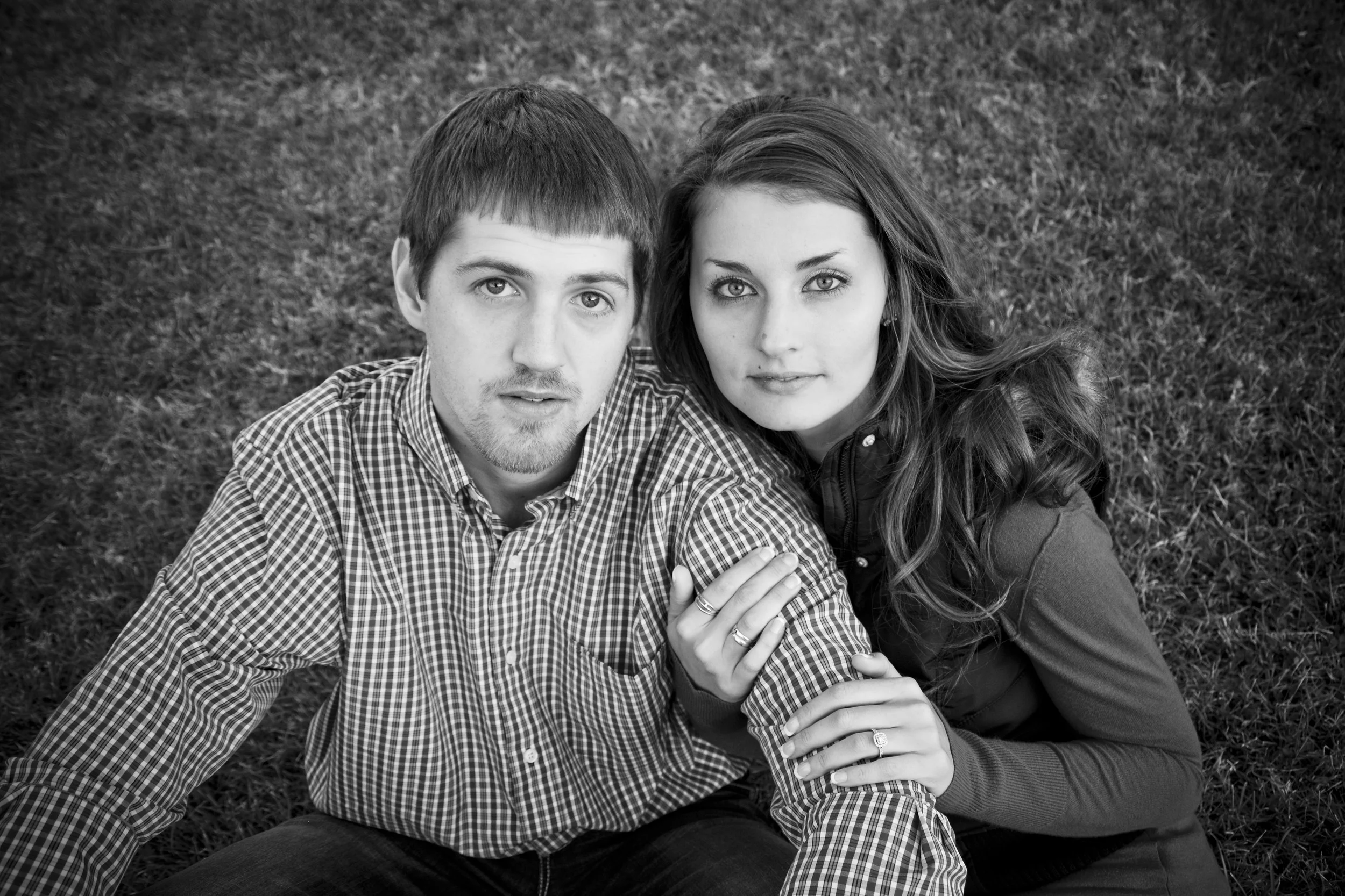 A black and white photo of a young man with short hair and a young woman with long hair, both lying on grass and looking directly at the camera.
