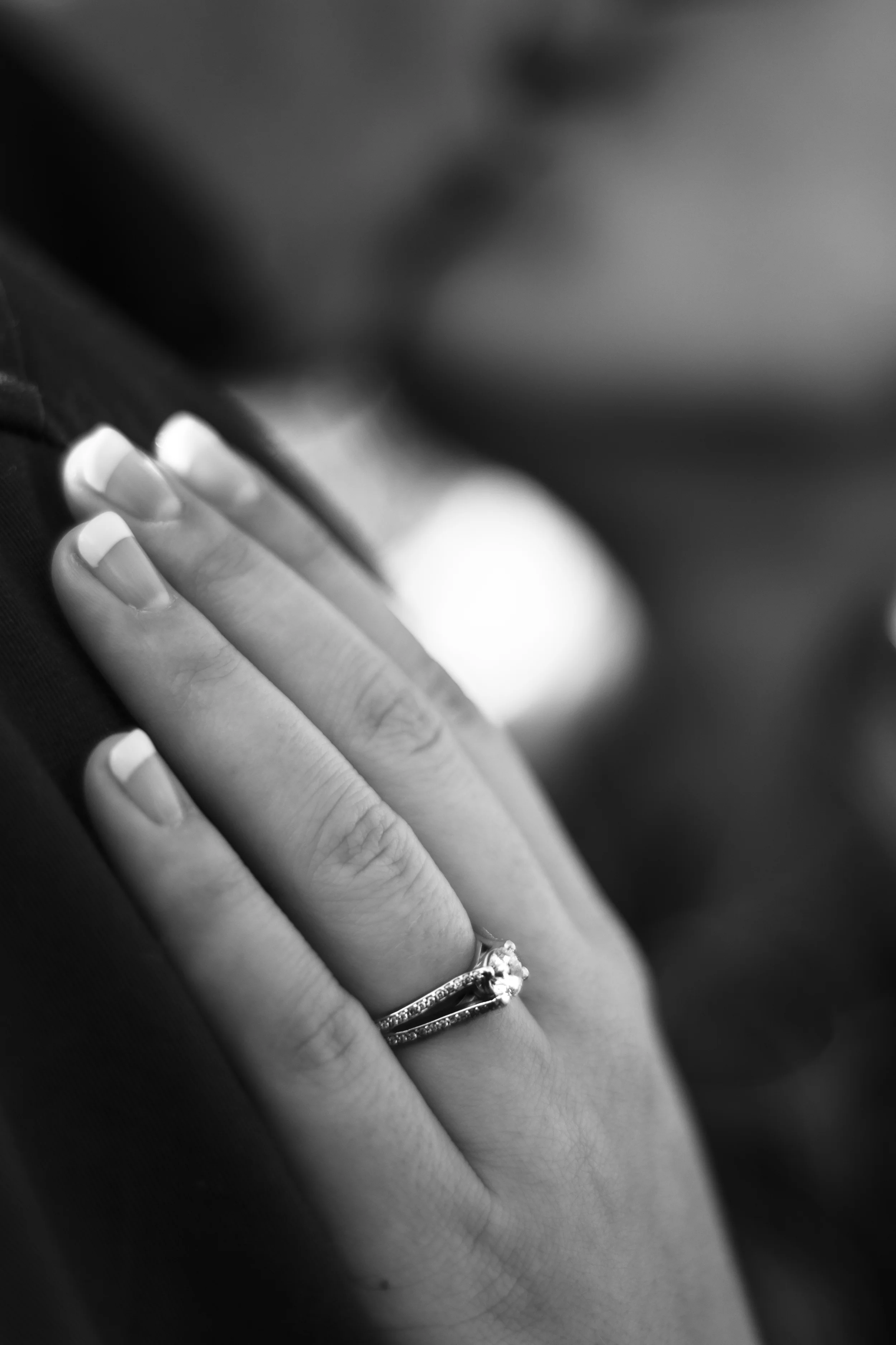 Close-up of a person's hand with a ring on the ring finger, showing white French manicure nails. The hand is resting on a dark surface, and the focus is on the fingers and rings.